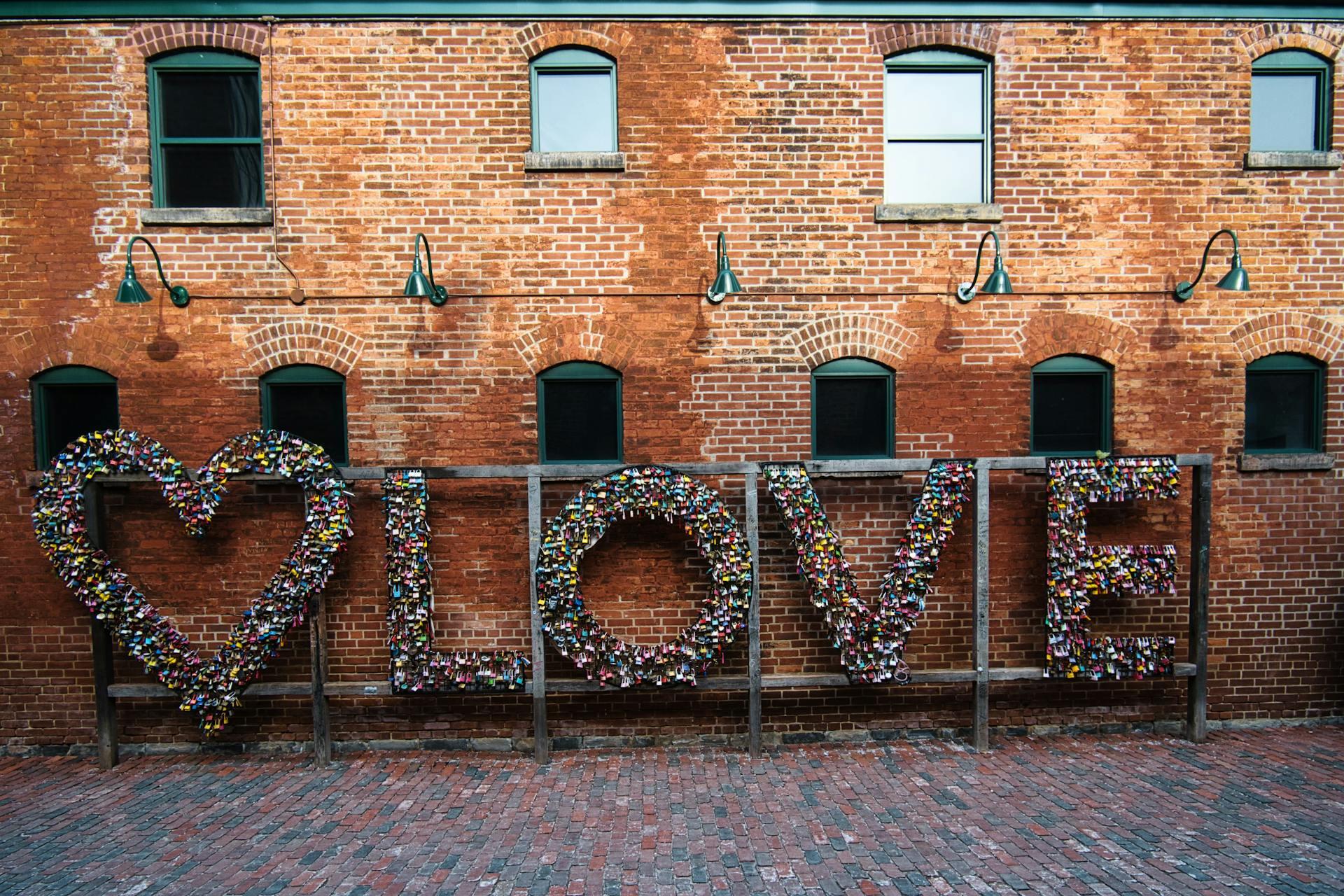 A brick wall features a large artistic display of "LOVE" made from colorful padlocks and a heart, creating a vibrant, romantic atmosphere.