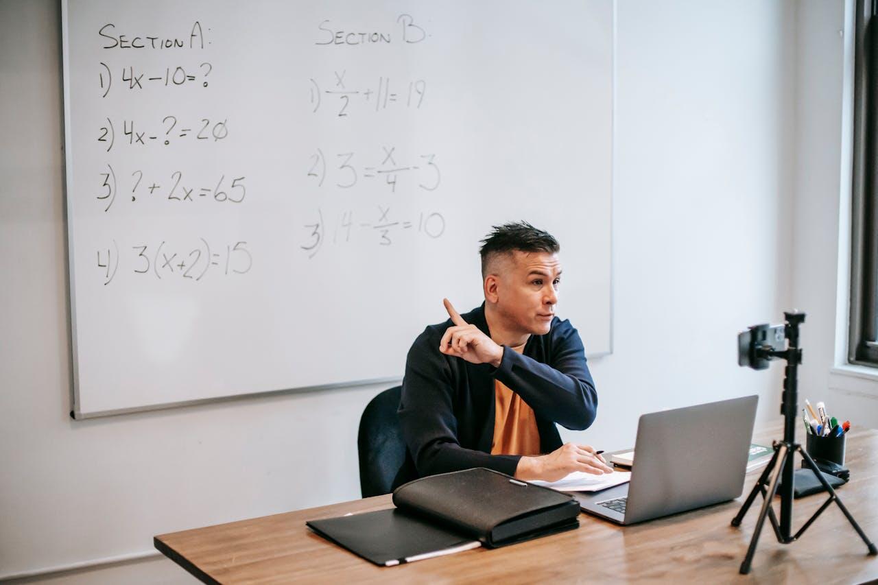 A person gestures while sitting at a desk with a laptop, surrounded by math equations on a whiteboard in a bright classroom.
