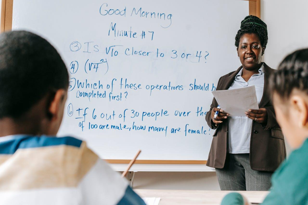 A teacher stands in front of a whiteboard with math problems, engaging with students seated at their desks.