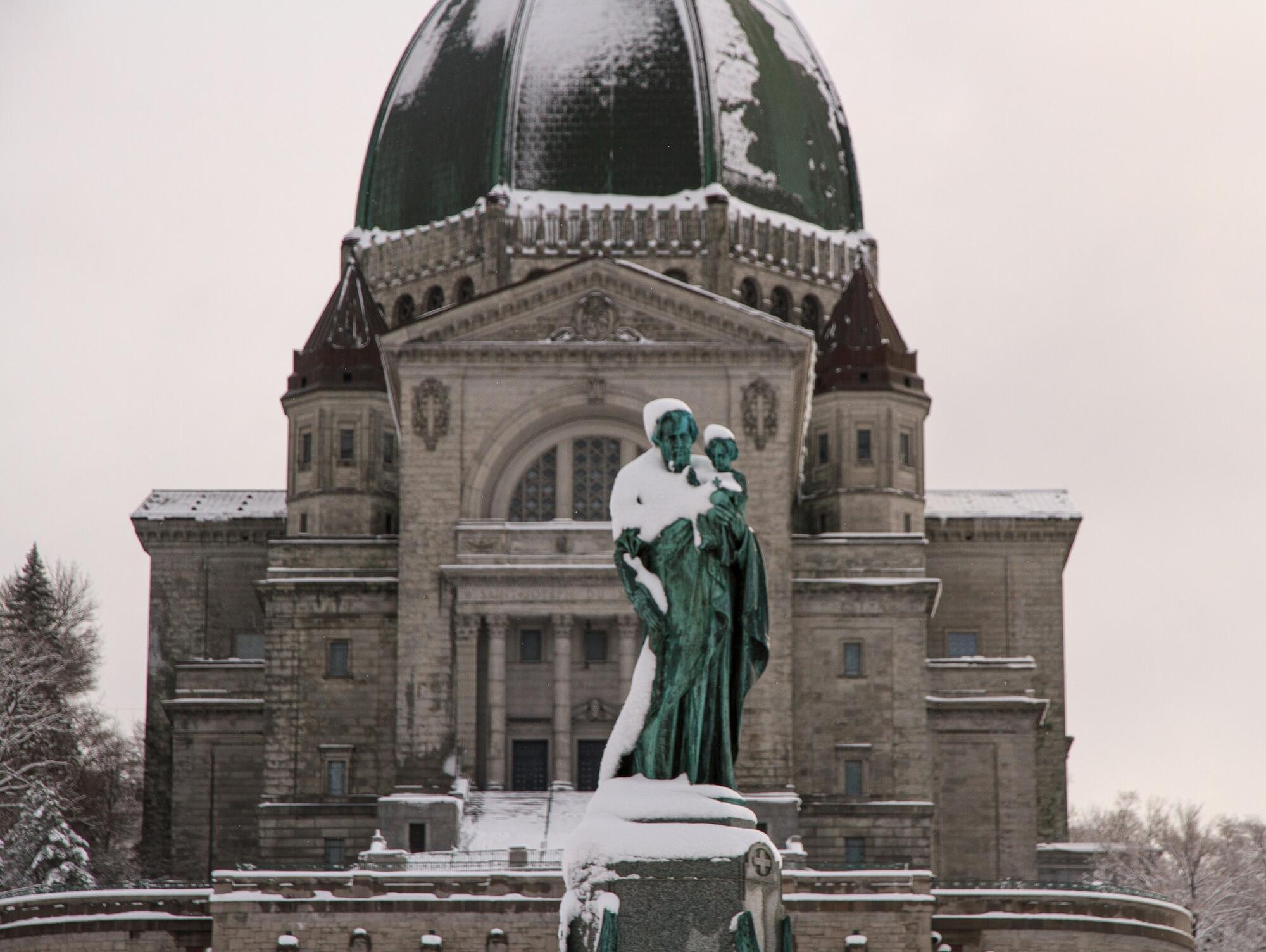 Statue in snow with a grand stone building in the background featuring a large, green dome and ornate architecture.