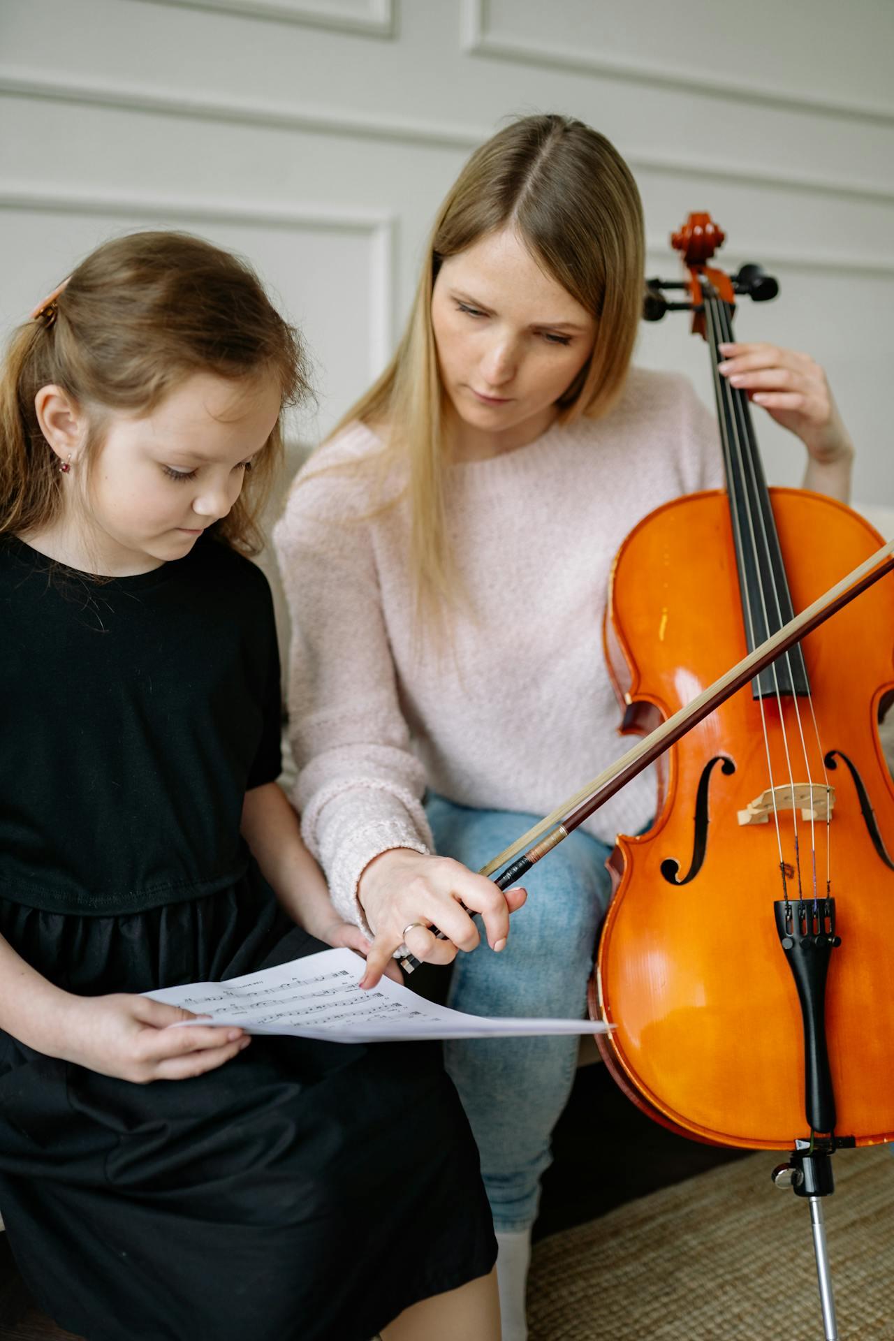 A woman teaches a girl to play the cello while referring to sheet music during a music lesson at home.