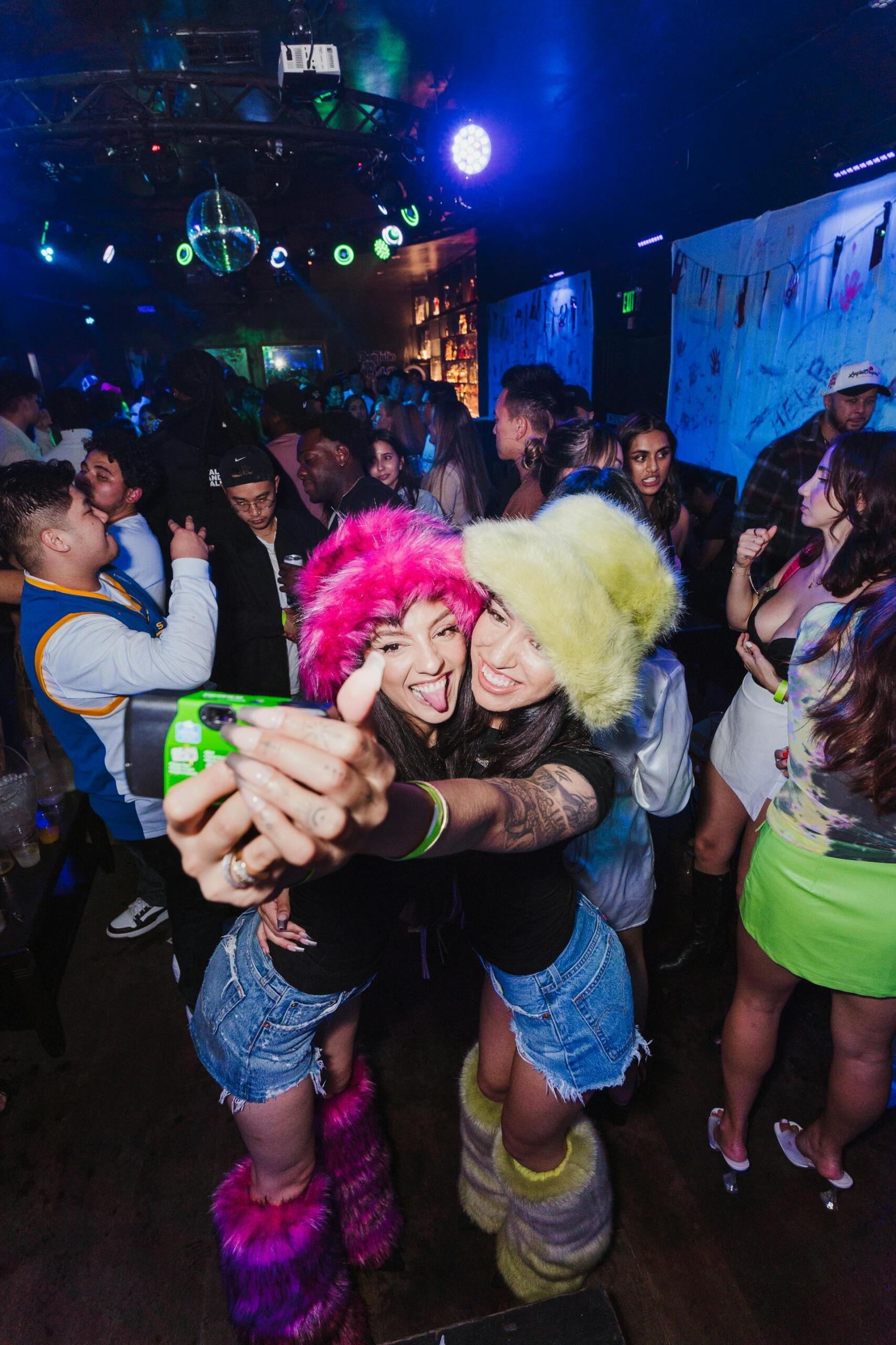 Two friends pose for a selfie at a lively party, surrounded by a colorful crowd and vibrant lighting.