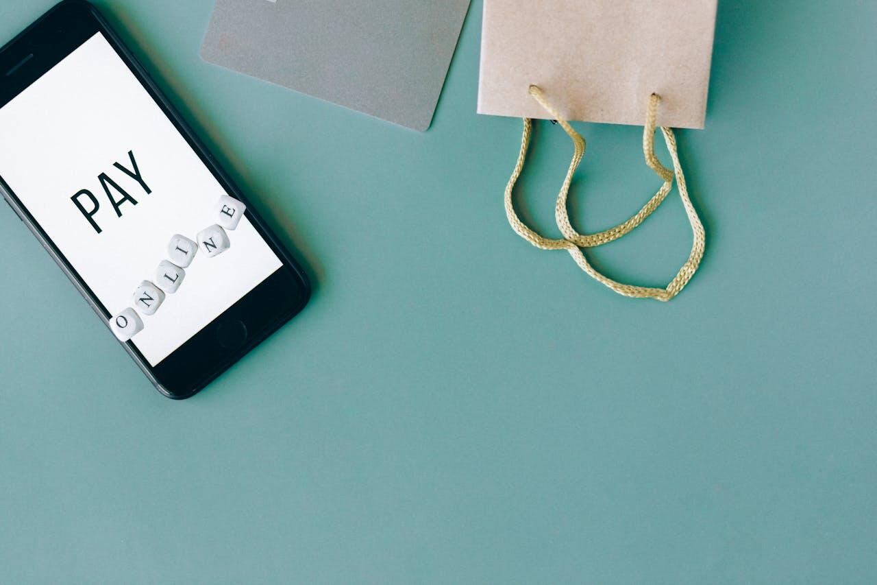 A smartphone displaying "PAY ONLINE" with letter beads nearby, next to a small brown shopping bag on a teal background.