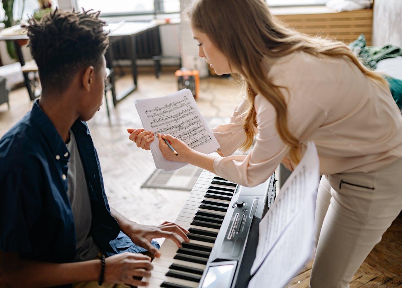 A young person plays a keyboard while a seated musician demonstrates music notes from a sheet, in a bright room filled with greenery.