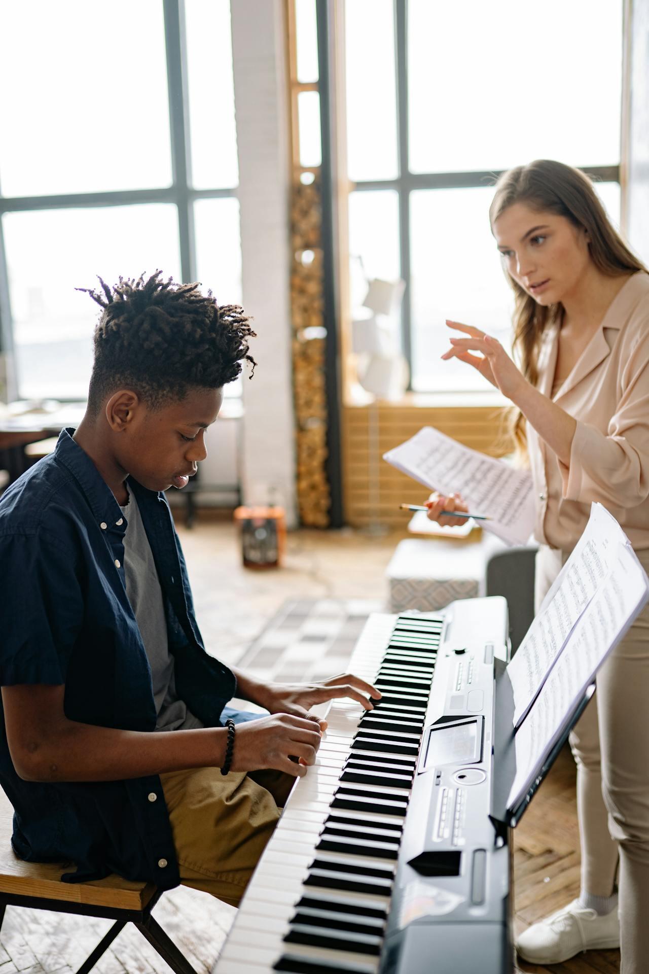 A young musician plays a keyboard while a mentor gestures and offers guidance, surrounded by a bright, creative space.