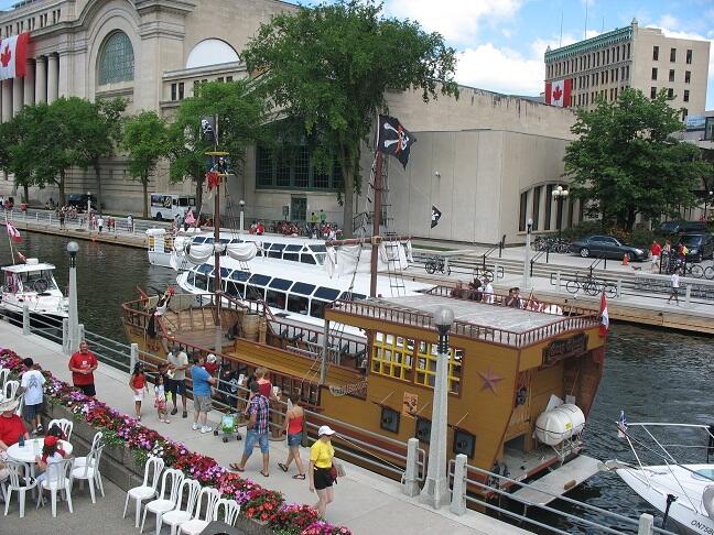 A pirate-themed boat docked on a canal, surrounded by people and colorful flowers, with buildings and trees in the background.
