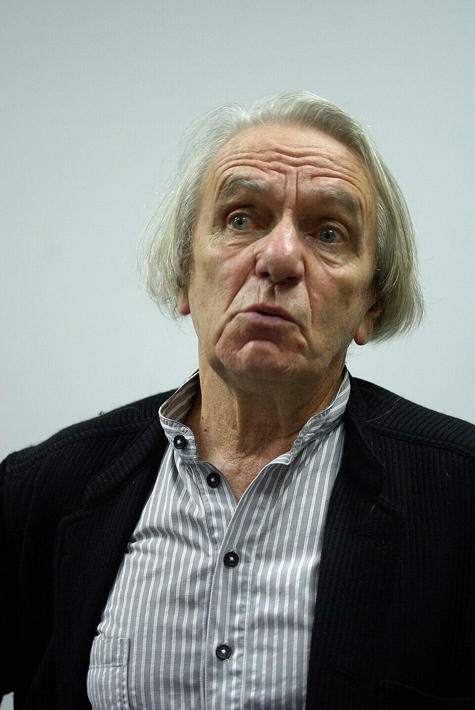 A color image of philosopher Jacques Rancière wearing a black jacket and a gray striped shirt speaking during an interview. 