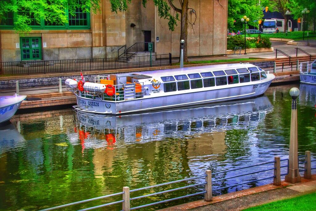 A silver boat named "Ottawa Queen" is docked in a calm canal, reflecting surrounding greenery and buildings.