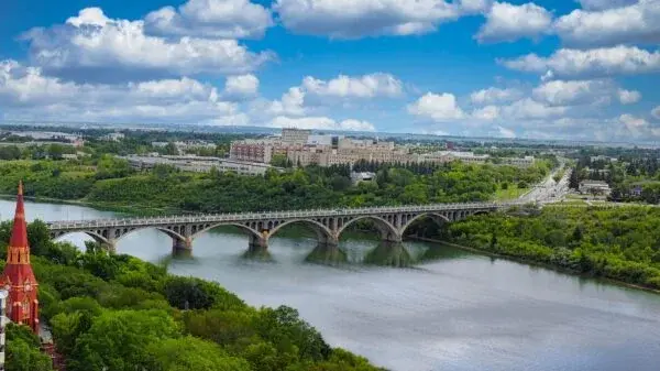 A scenic view of a large bridge over a river surrounded by lush greenery and modern buildings under a partly cloudy sky.