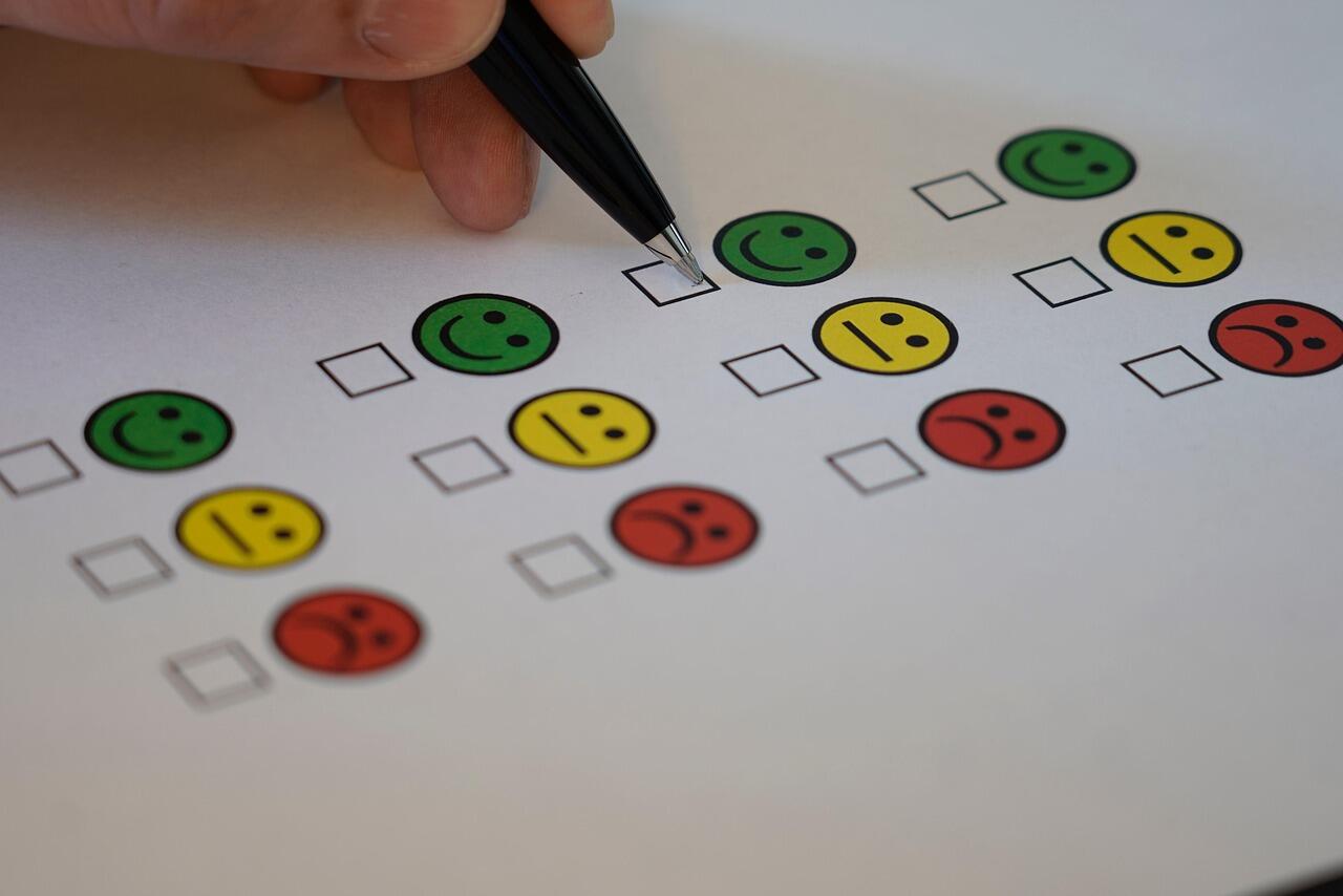 A hand holds a pen above a printed feedback form with green, yellow, and red smiley faces and checkboxes for responses.
