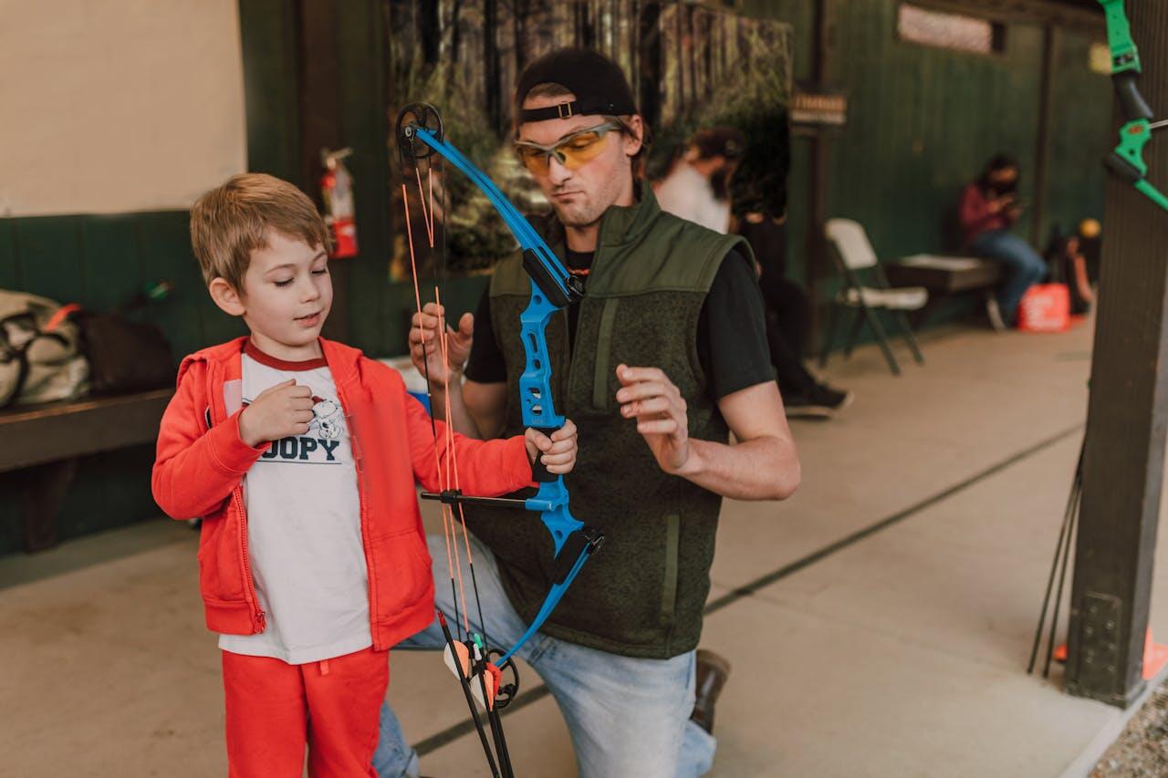 A child in a red outfit holds a blue bow, while an adult guides them, both smiling in an archery setting. Chairs and bags in the background.
