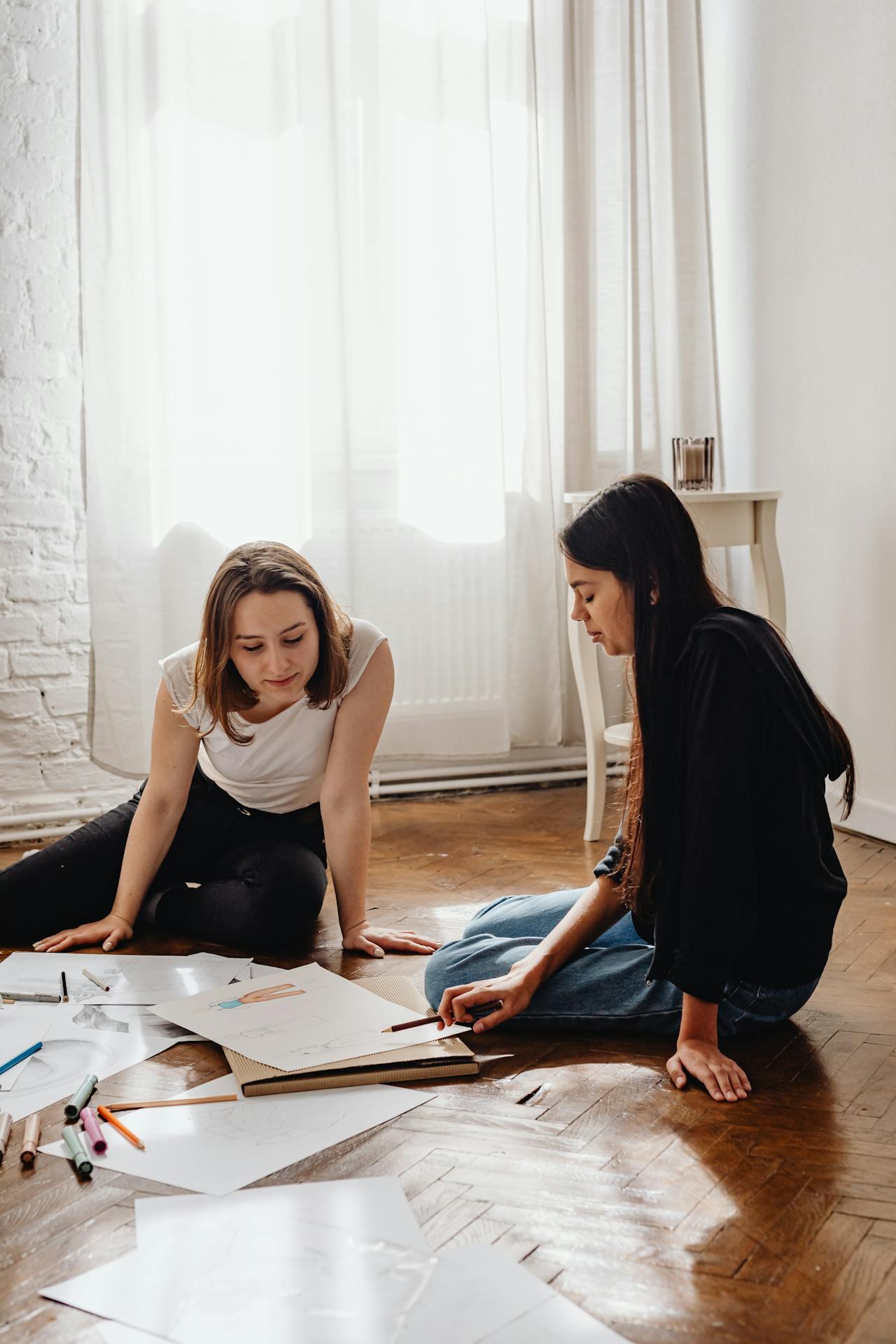 Two individuals sit on the floor surrounded by scattered papers and art supplies, engaged in a creative discussion.