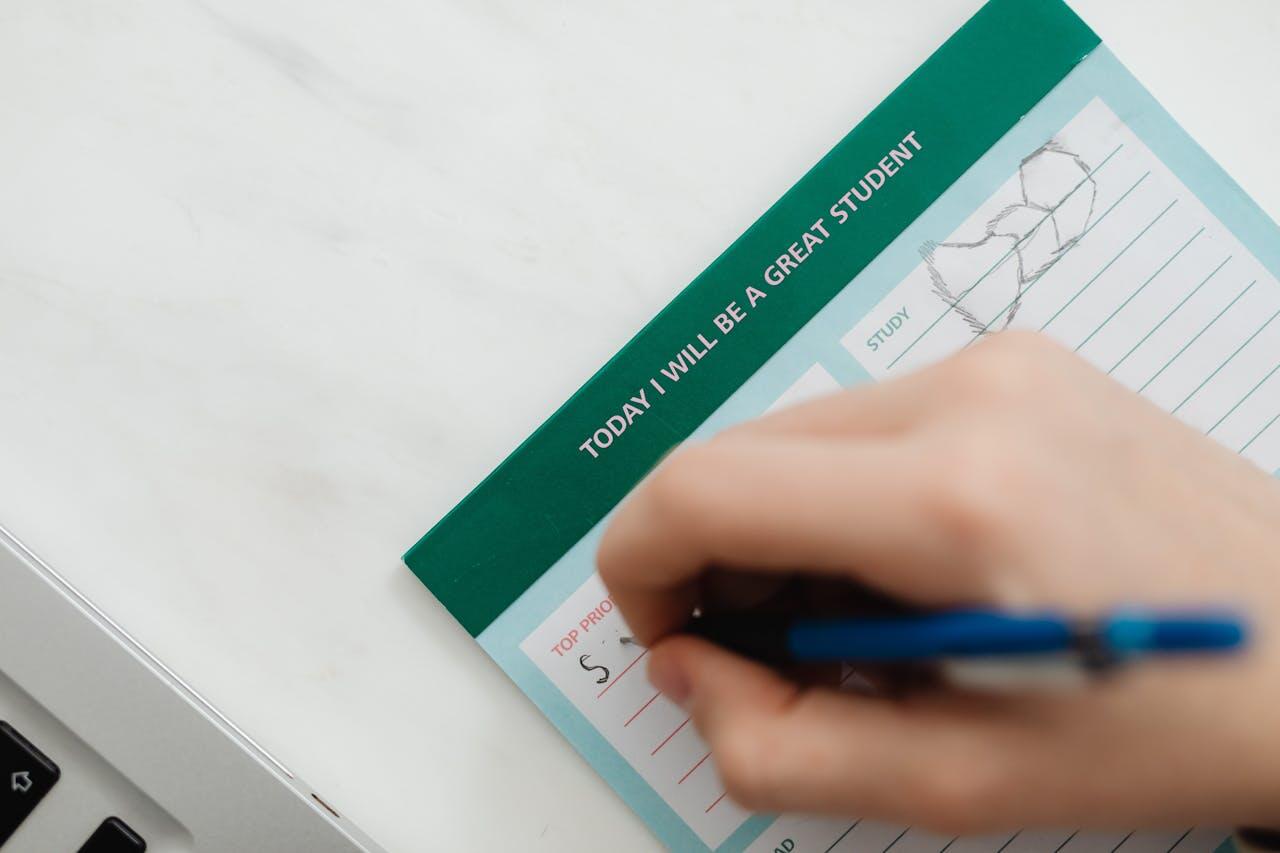 A close-up of a hand writing on a teal and green planner page, which reads, "Today I will be a great student." A laptop is visible in the background.