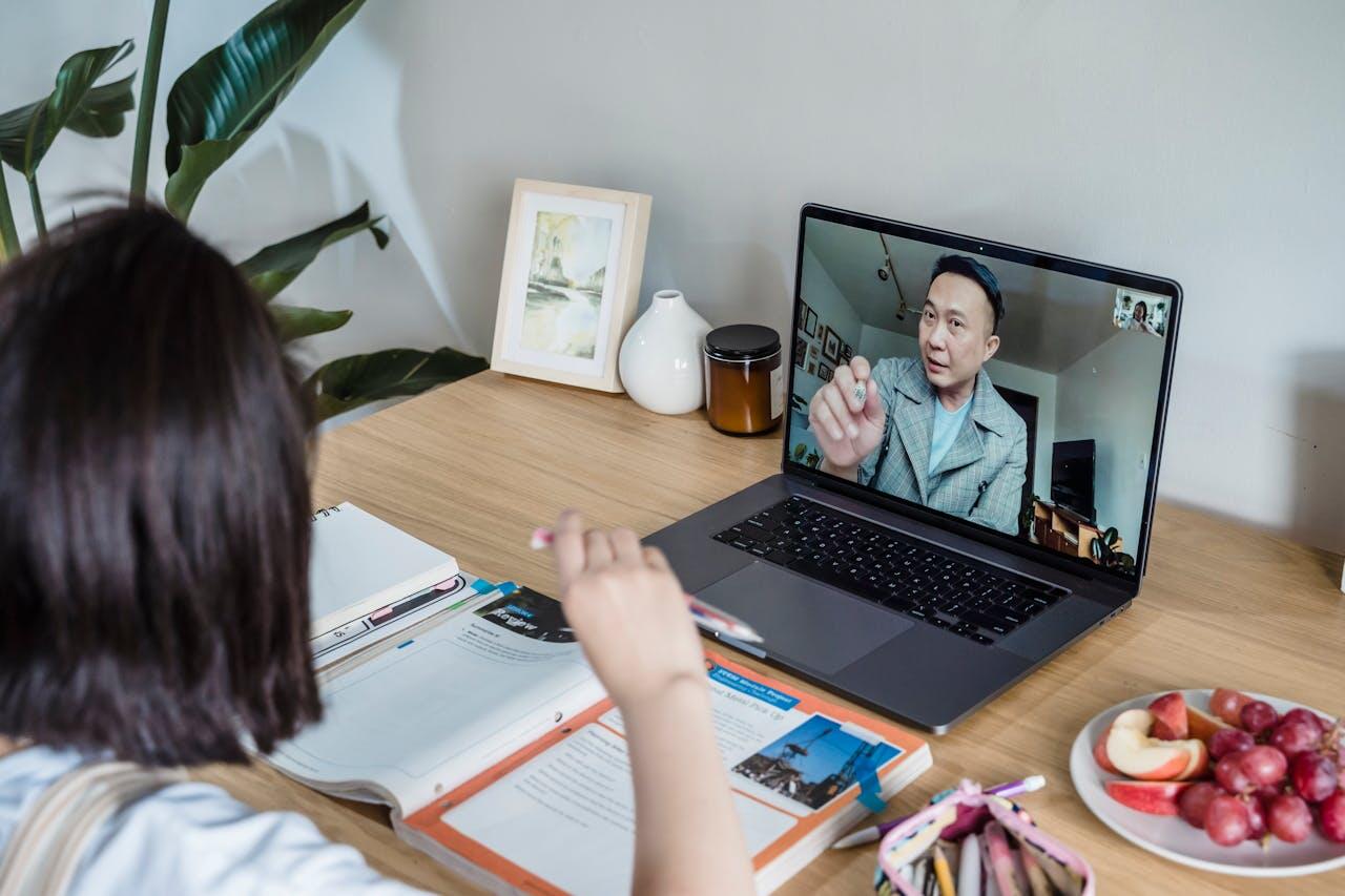 A person is attending a video call on a laptop while studying at a wooden desk, surrounded by books, pens, and a plate of fruit.