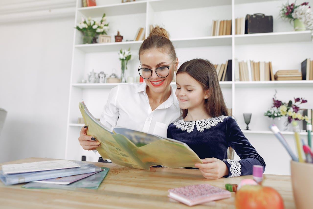 A woman and a girl sit together at a table, reading a colorful book, surrounded by school supplies and decorative items.