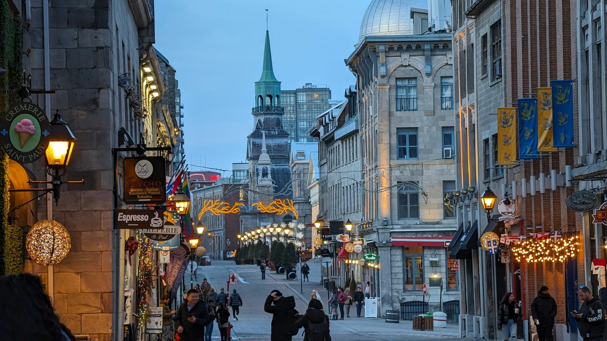 A charming street in a historic area, illuminated by warm lights, featuring shops, cafés, and a view of a distant steeple against a twilight sky.
