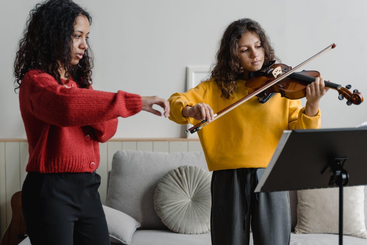 A child in a yellow sweater plays violin while an adult in a red sweater gestures, guiding them in a cozy indoor space.