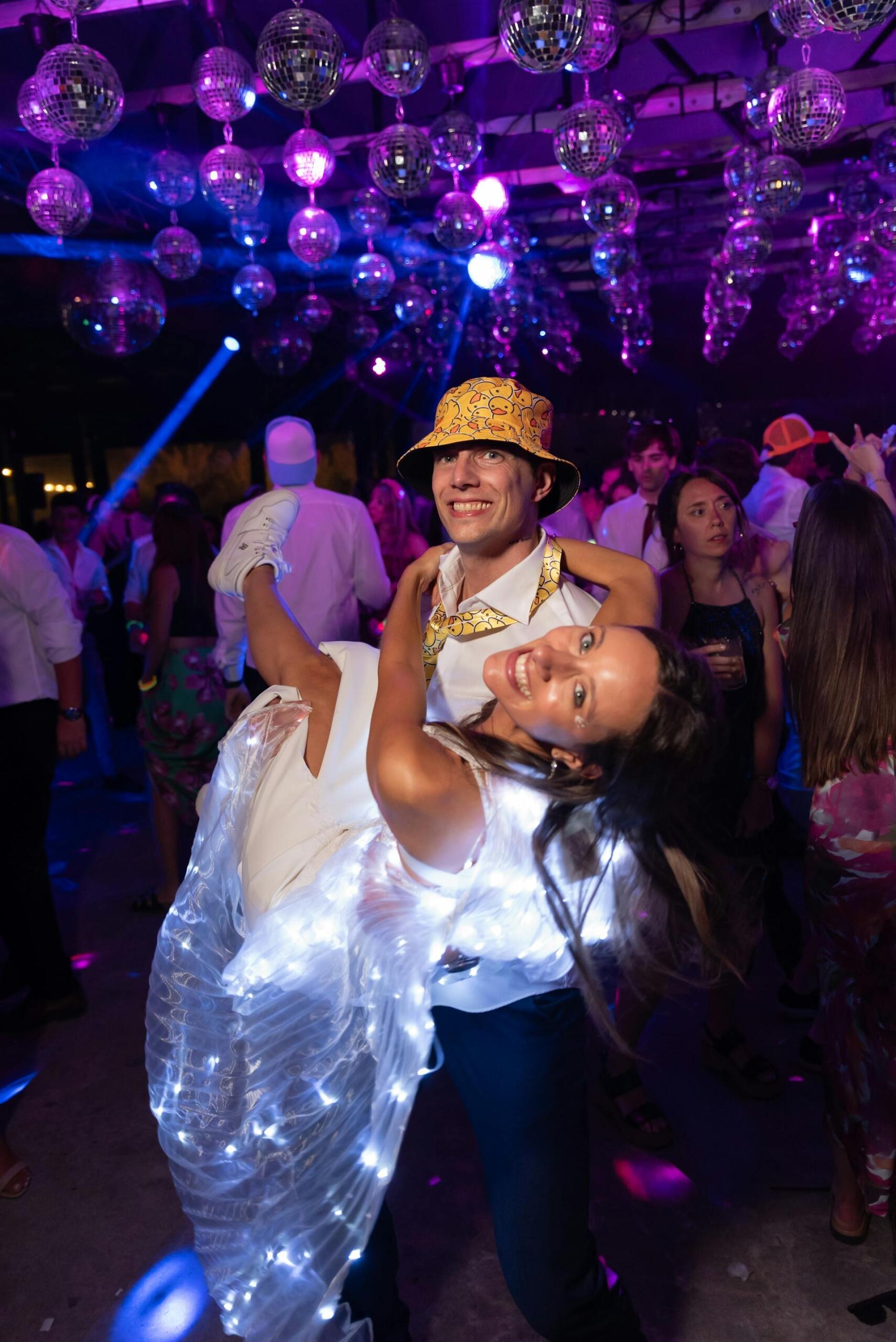 A couple dances joyfully at a vibrant party, with disco balls above and colorful lights illuminating their festive outfits.