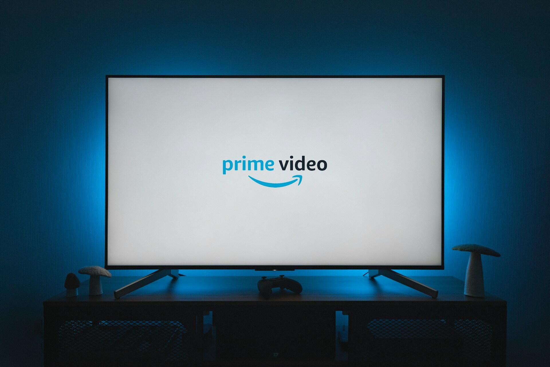 A TV displaying the Prime Video logo, illuminated by blue backlighting, with gaming controller and decorative mushrooms on a dark wooden stand.