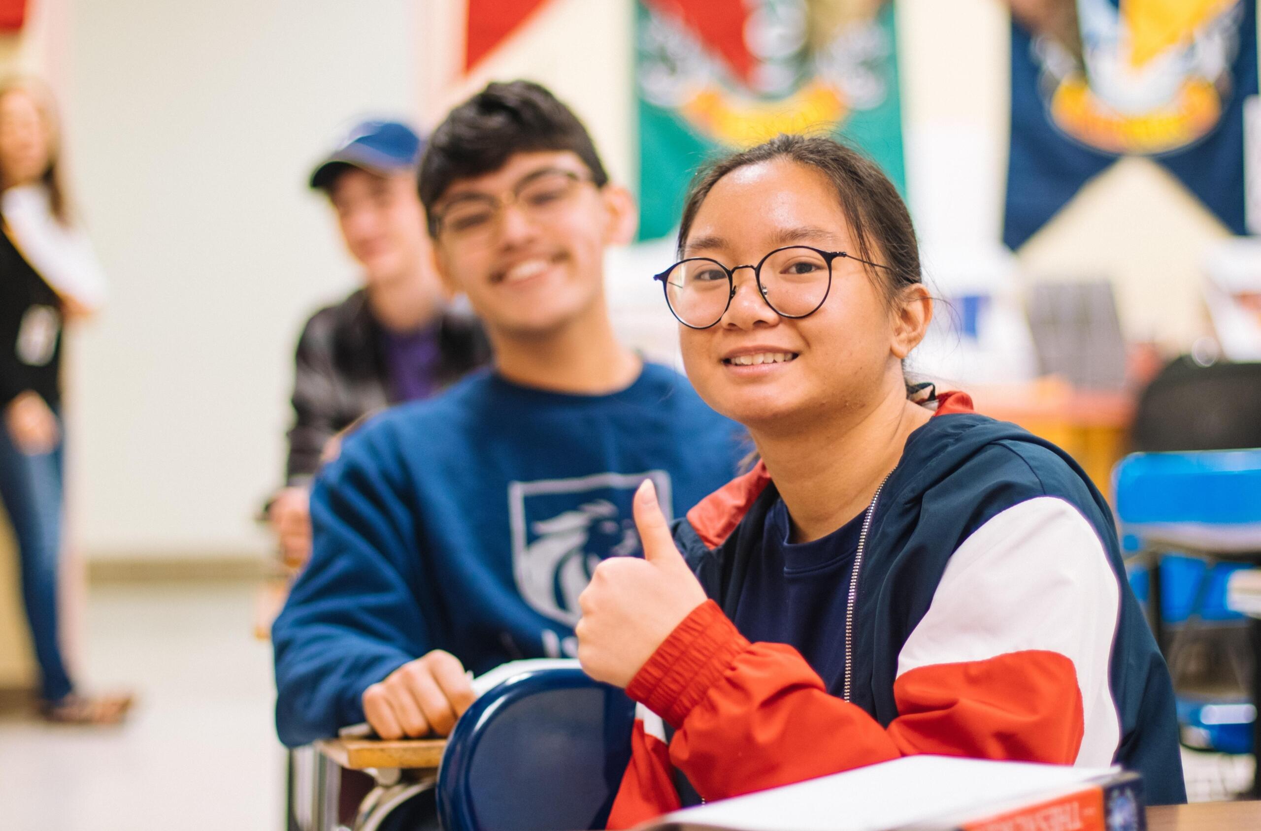 High school students sitting in a classroom, smiling at the camera, with one student giving a thumbs-up while seated at a desk.