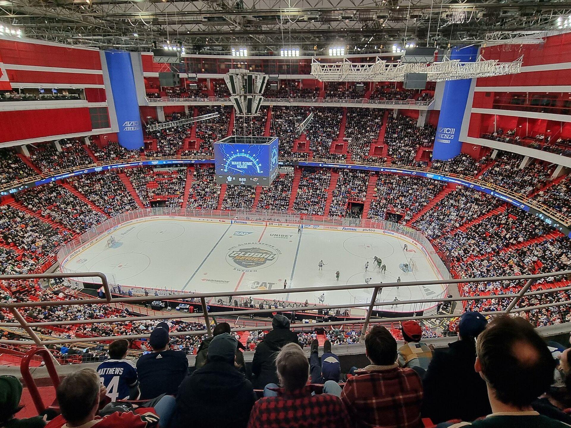 Aerial view of a packed hockey arena with fans watching a game on the ice, illuminated by bright lights and a large scoreboard.