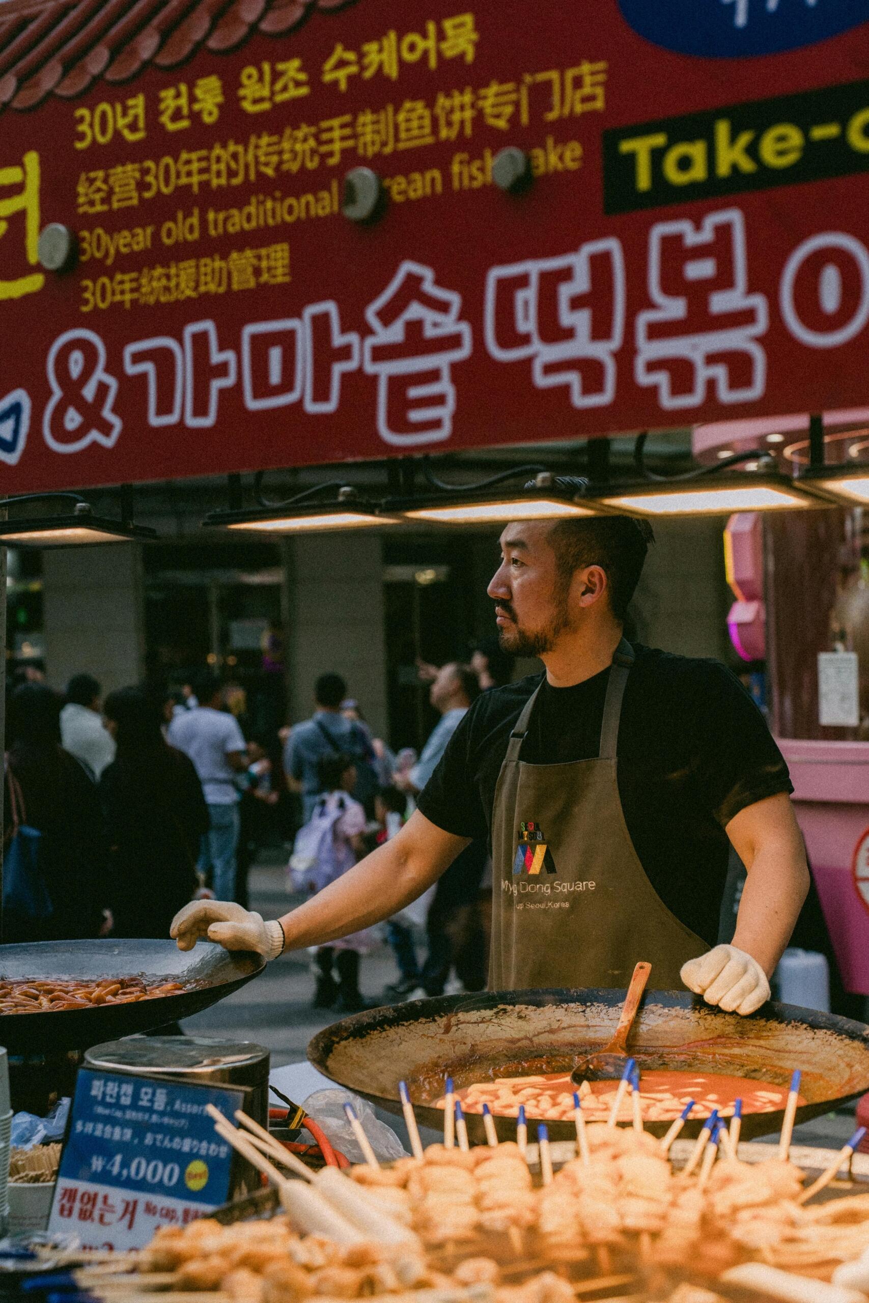 Korean man standing at a street food stall, preparing or serving food during a cultural festival.