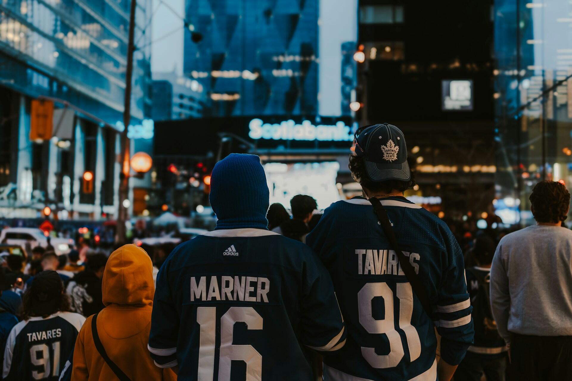 Two fans in Toronto Maple Leafs jerseys watch a city scene at night, engrossed in the excitement around them.