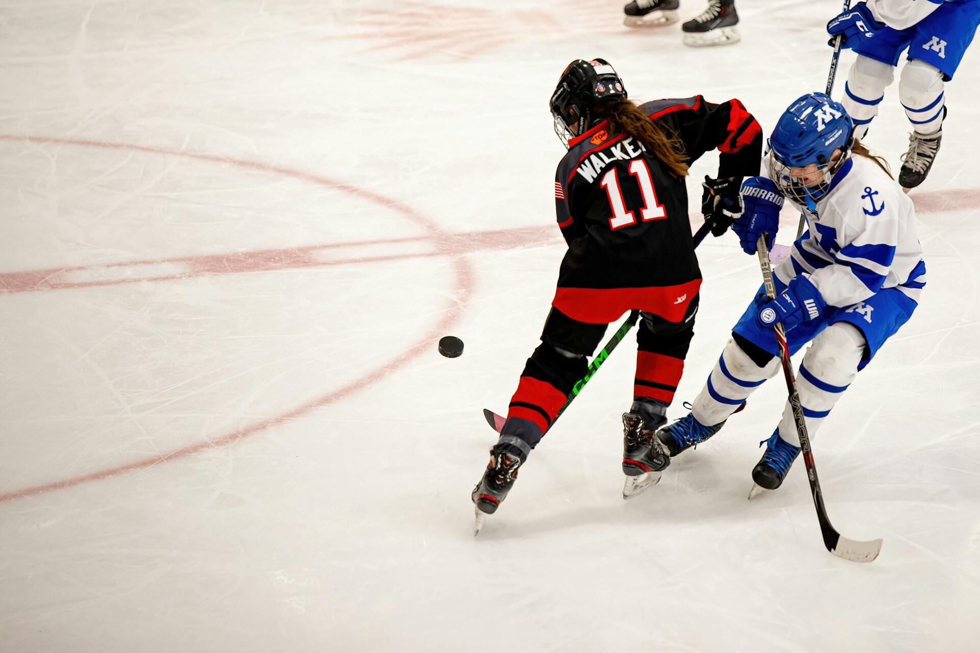 A female hockey player in a black and red uniform competes for the puck against another player in blue and white on the ice.