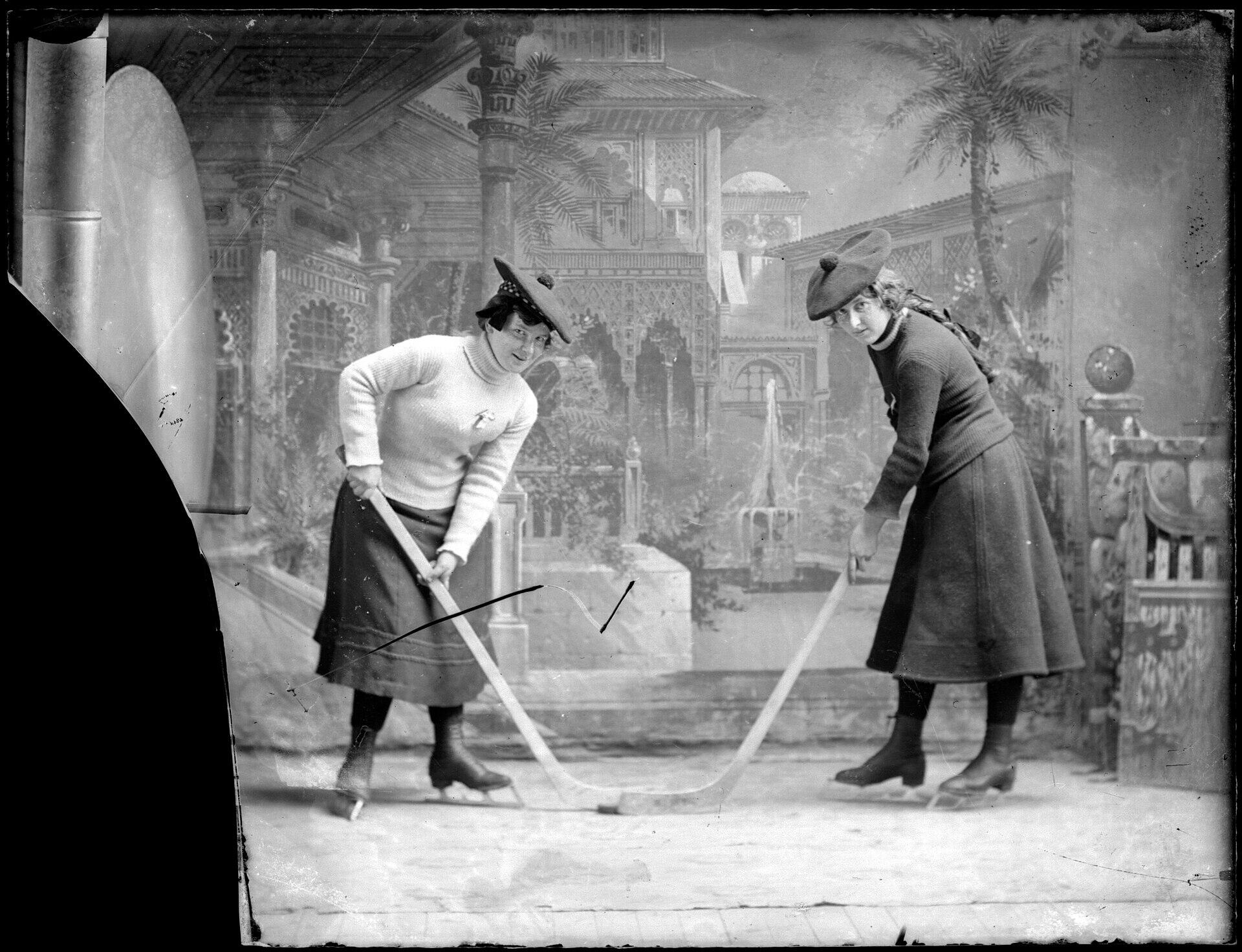 Two women dressed in early 20th-century fashion play curling on a decorated stage with a backdrop of ornate architecture.