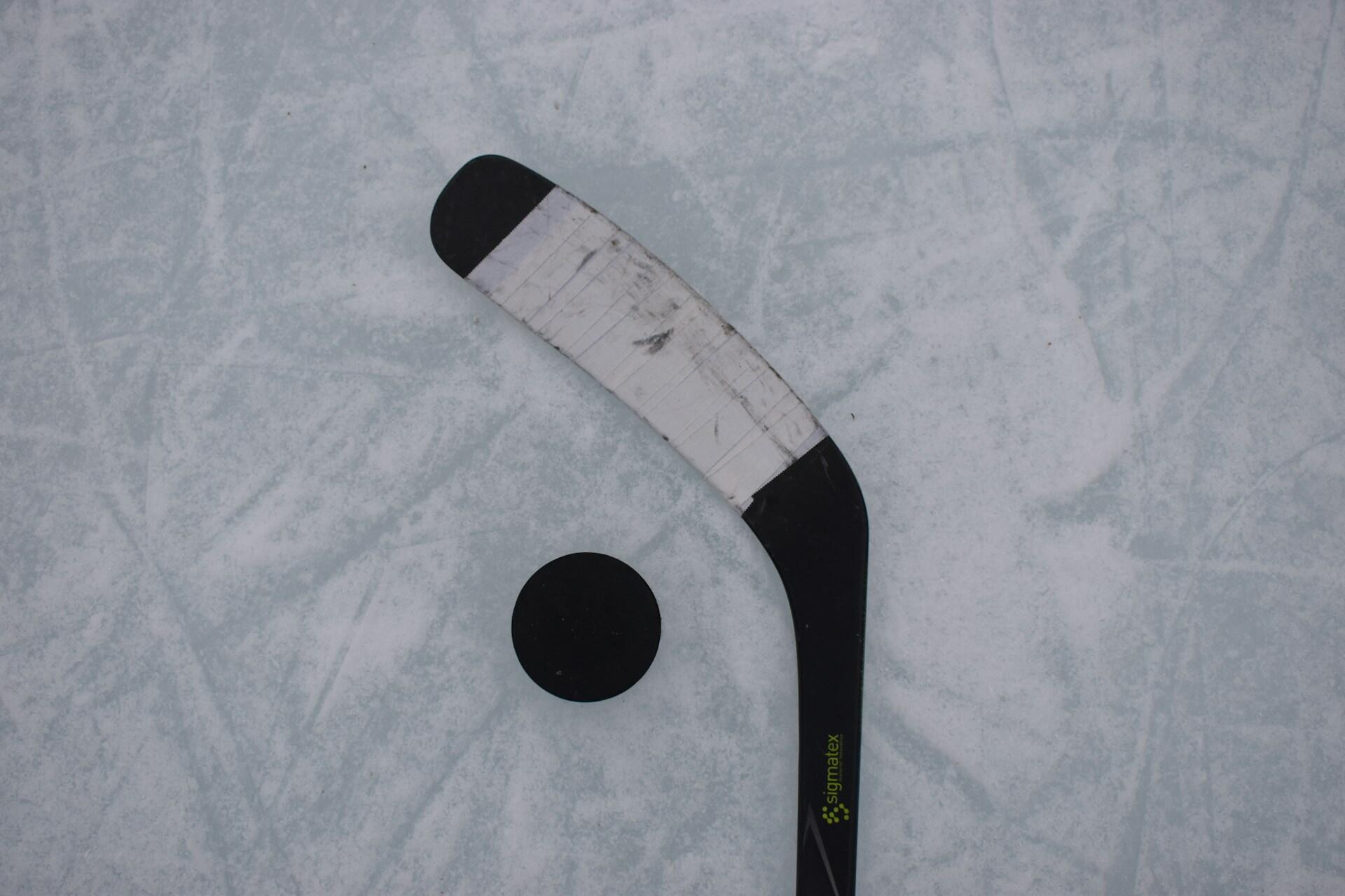 A close-up view of a hockey stick and a black puck on an icy surface, showcasing the stick's curved blade and scratched finish.