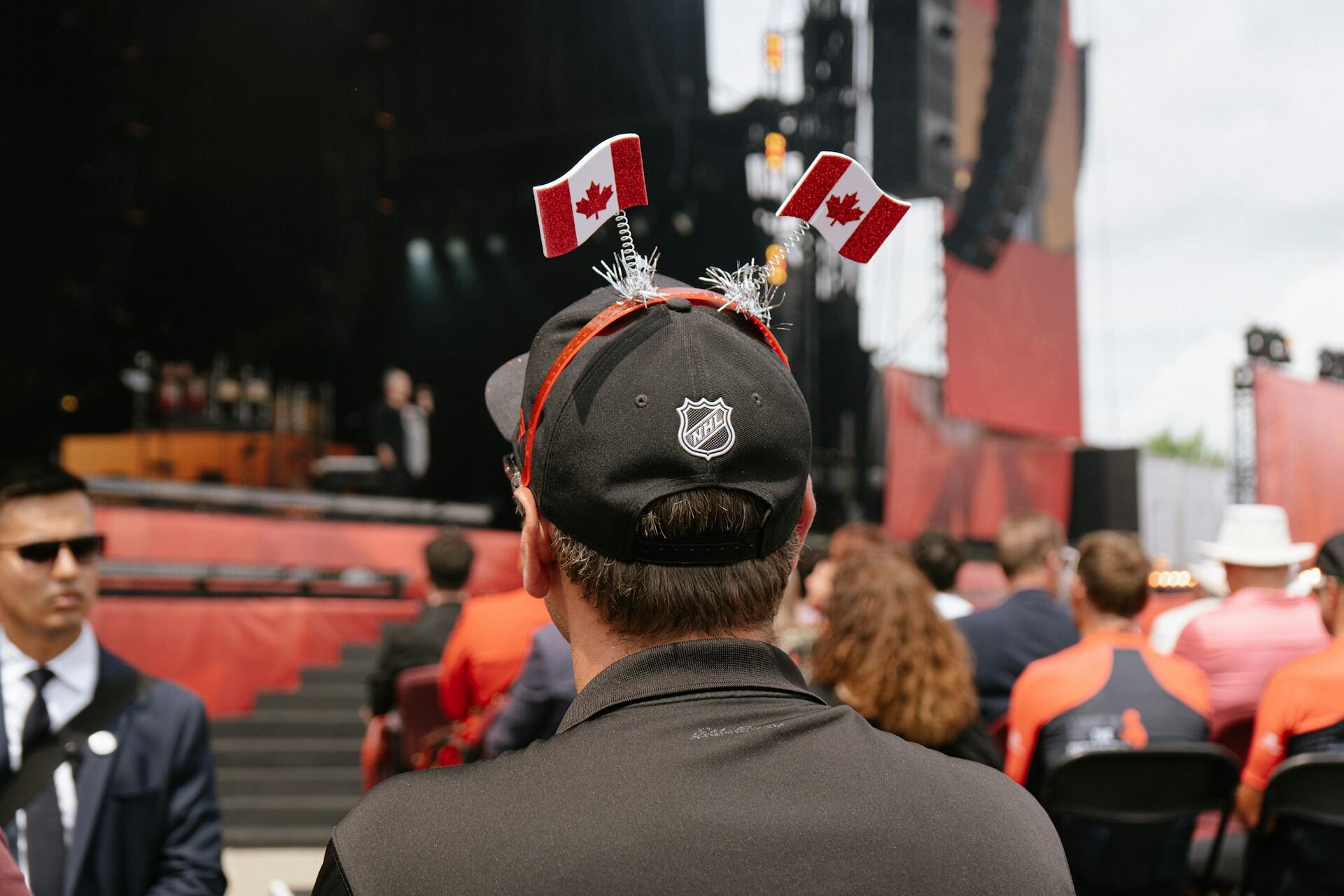 A person wearing a black cap with NHL logo and festive Canadian flags attends an outdoor event filled with spectators.