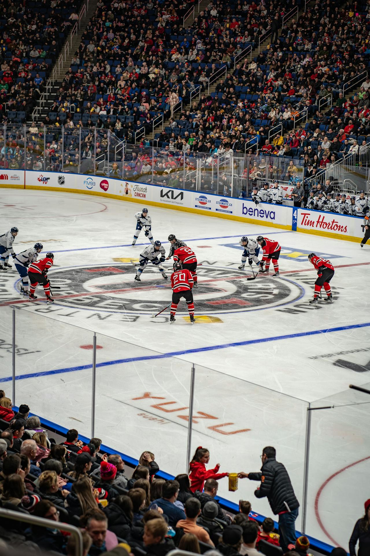 Hockey players face off at center ice during a game, with a packed arena of spectators in the background.