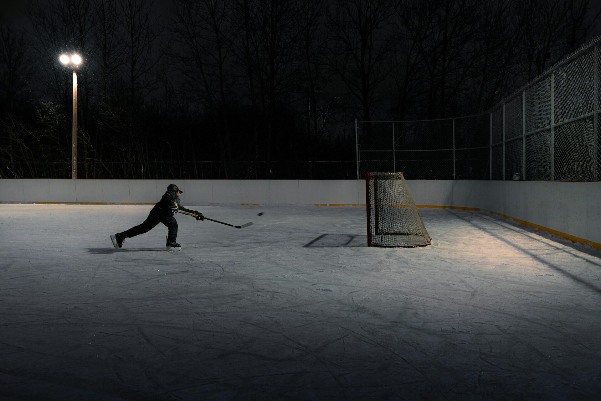 A lone hockey player shoots a puck toward an empty net on an illuminated outdoor rink, surrounded by darkness and trees.