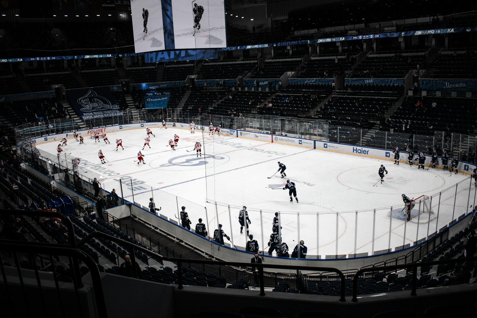 A hockey rink with players in red and blue jerseys warming up, surrounded by empty seats in a modern arena.