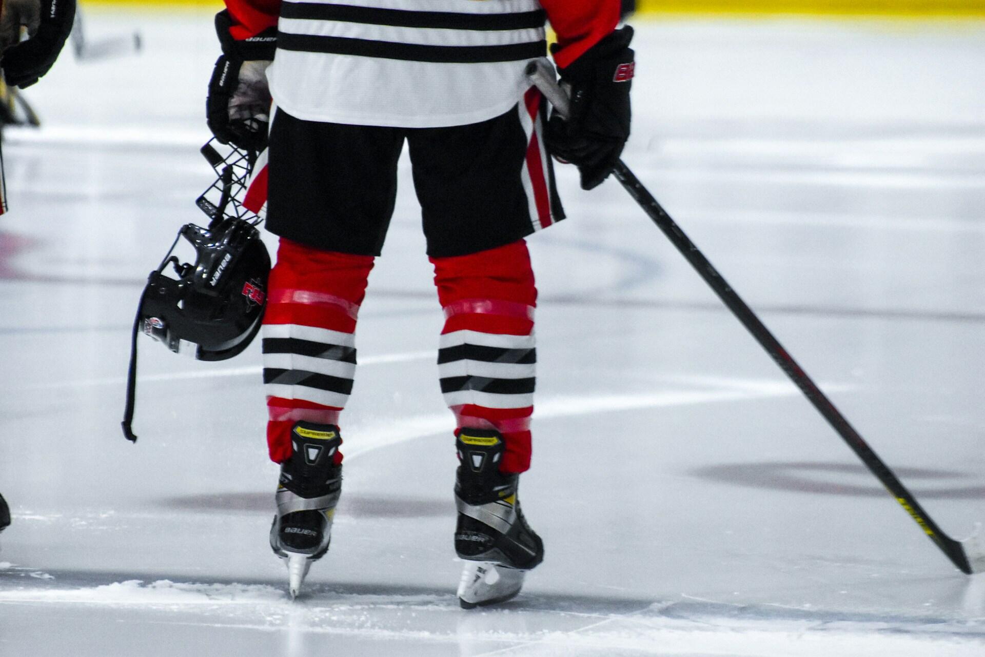 A hockey player stands on the ice, wearing red and black gear, holding a helmet in one hand and a stick in the other.