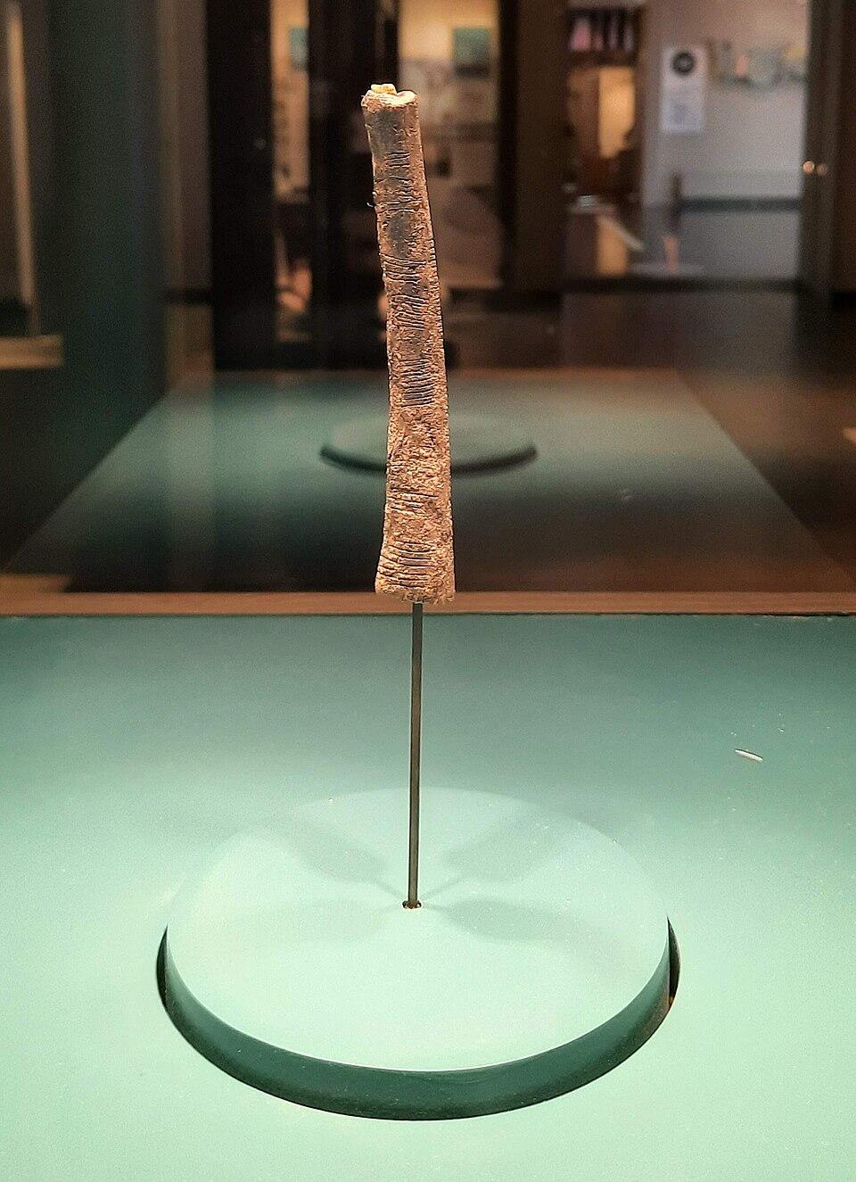 A slender, ancient artifact displayed upright on a metal stand, surrounded by a minimalist exhibit setting.