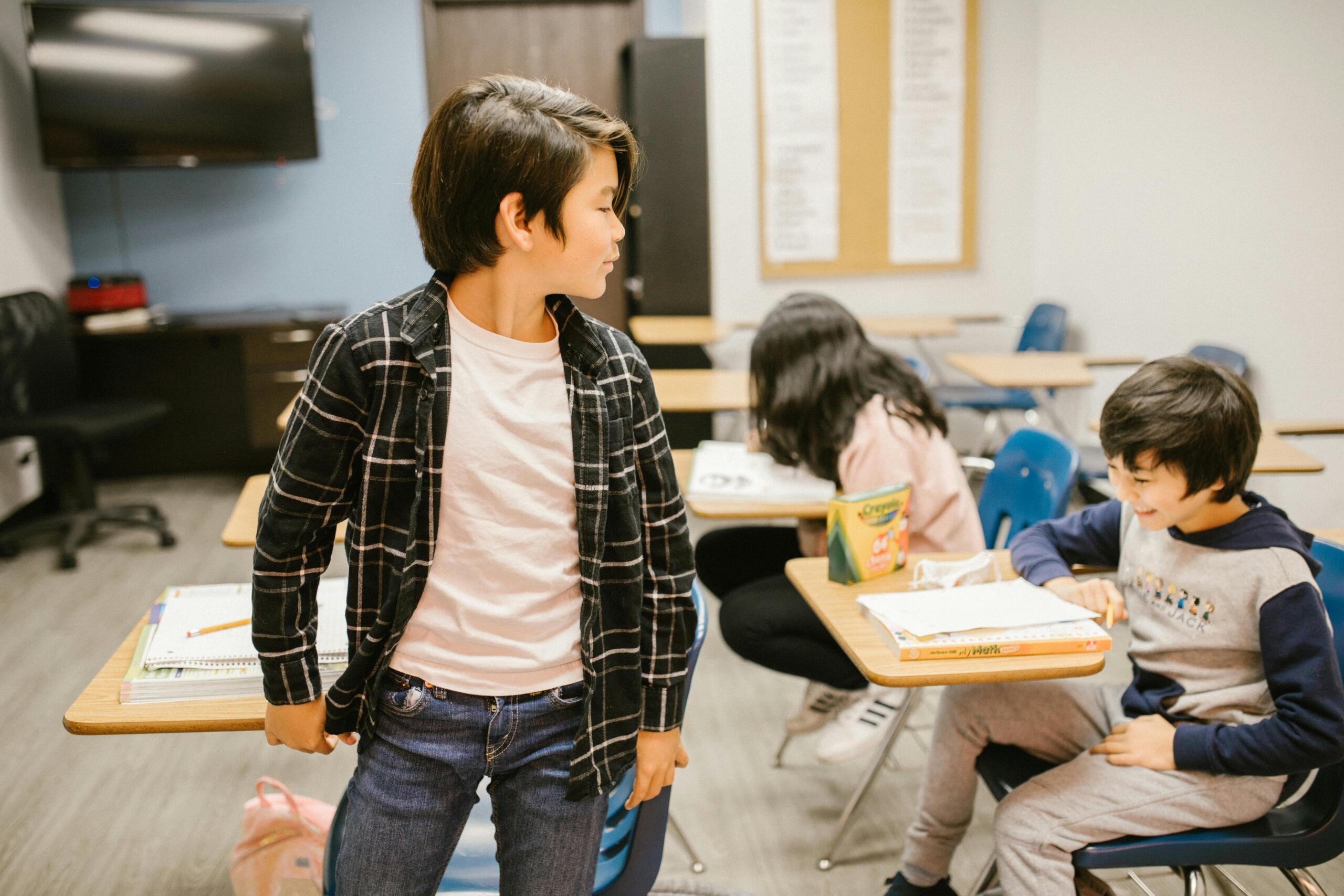Classroom of Japanese students sitting at desks while one student stands to speak, with classmates listening and a teacher nearby in a bright school setting.