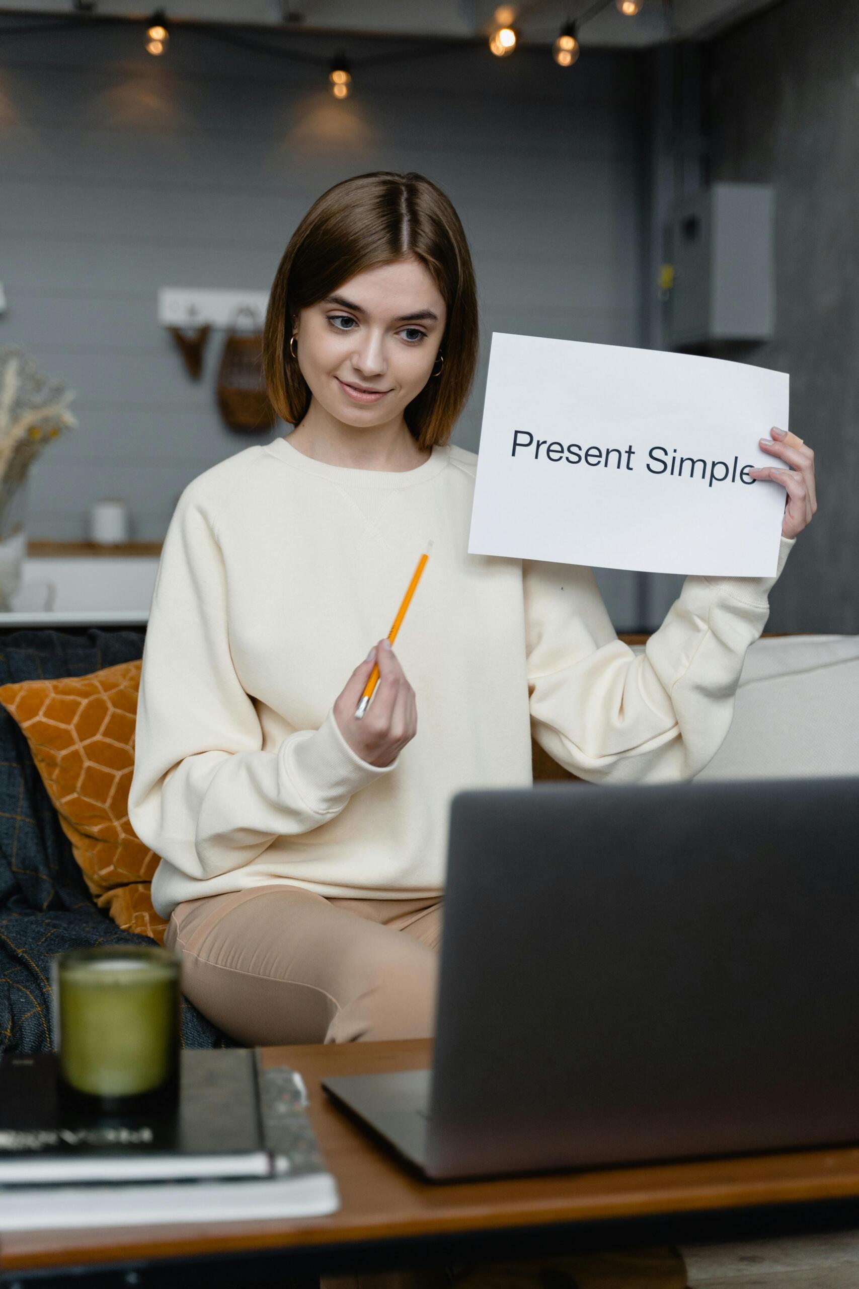 English teacher standing at the front of a classroom holding a sign that says “Present Simple,” with a whiteboard behind them and a teaching environment visible. Source: Pexels.