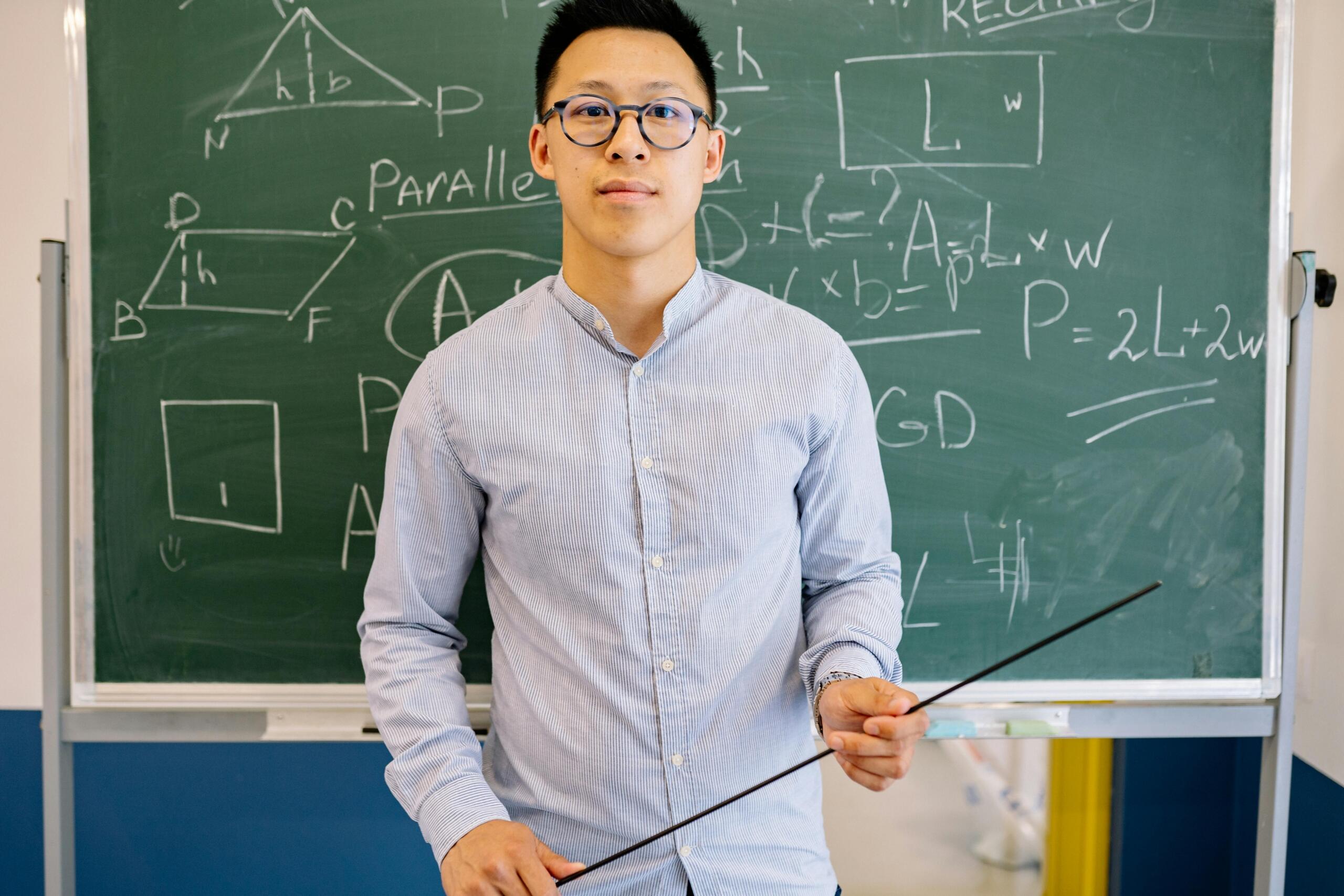 Teacher standing in front of a blackboard covered with mathematical formulas, explaining the lesson in a classroom setting.