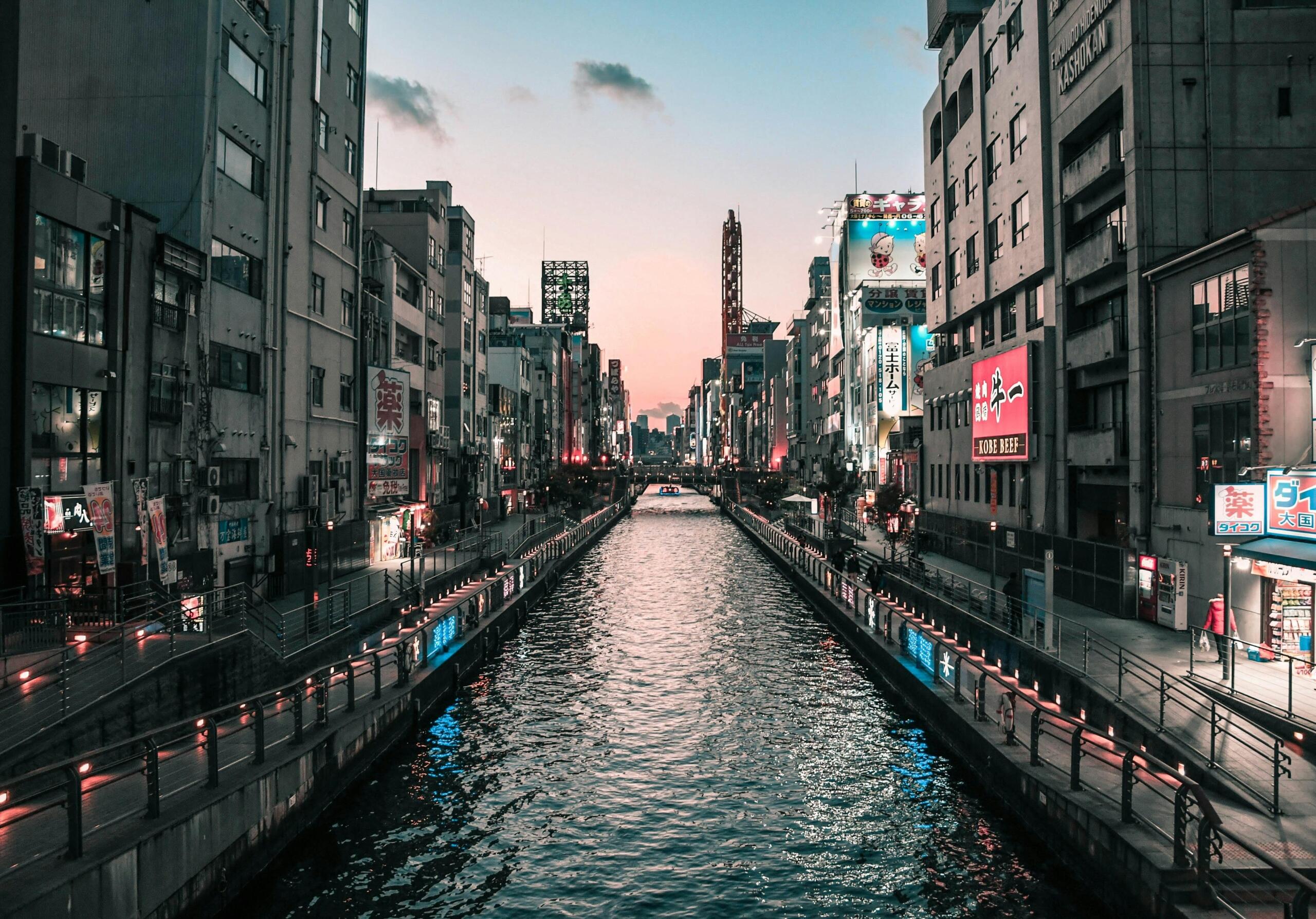 Twilight view of the Osaka River flowing through the center of the city, with illuminated skyscrapers and office buildings reflecting on the calm water, and a soft purple-blue evening sky overhead.Source: Satoshi.