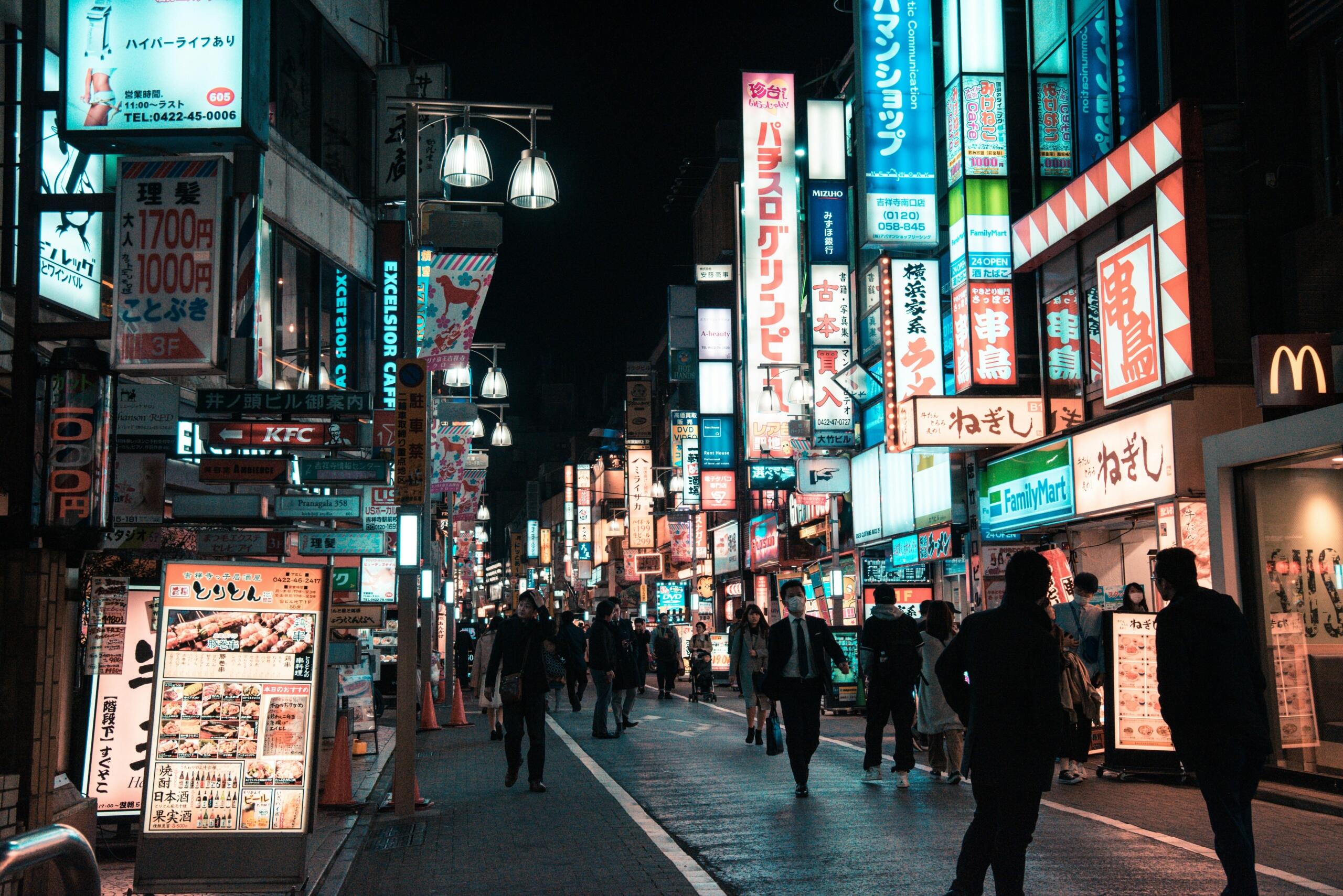 Busy Tokyo street at night illuminated by bright neon signs and storefront lights, with pedestrians walking along the sidewalks and cars passing through the glowing city scene.