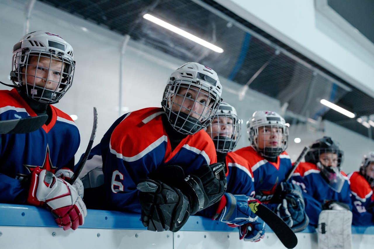 A group of young hockey players in blue and red uniforms lean against the rink boards, eagerly watching the game.