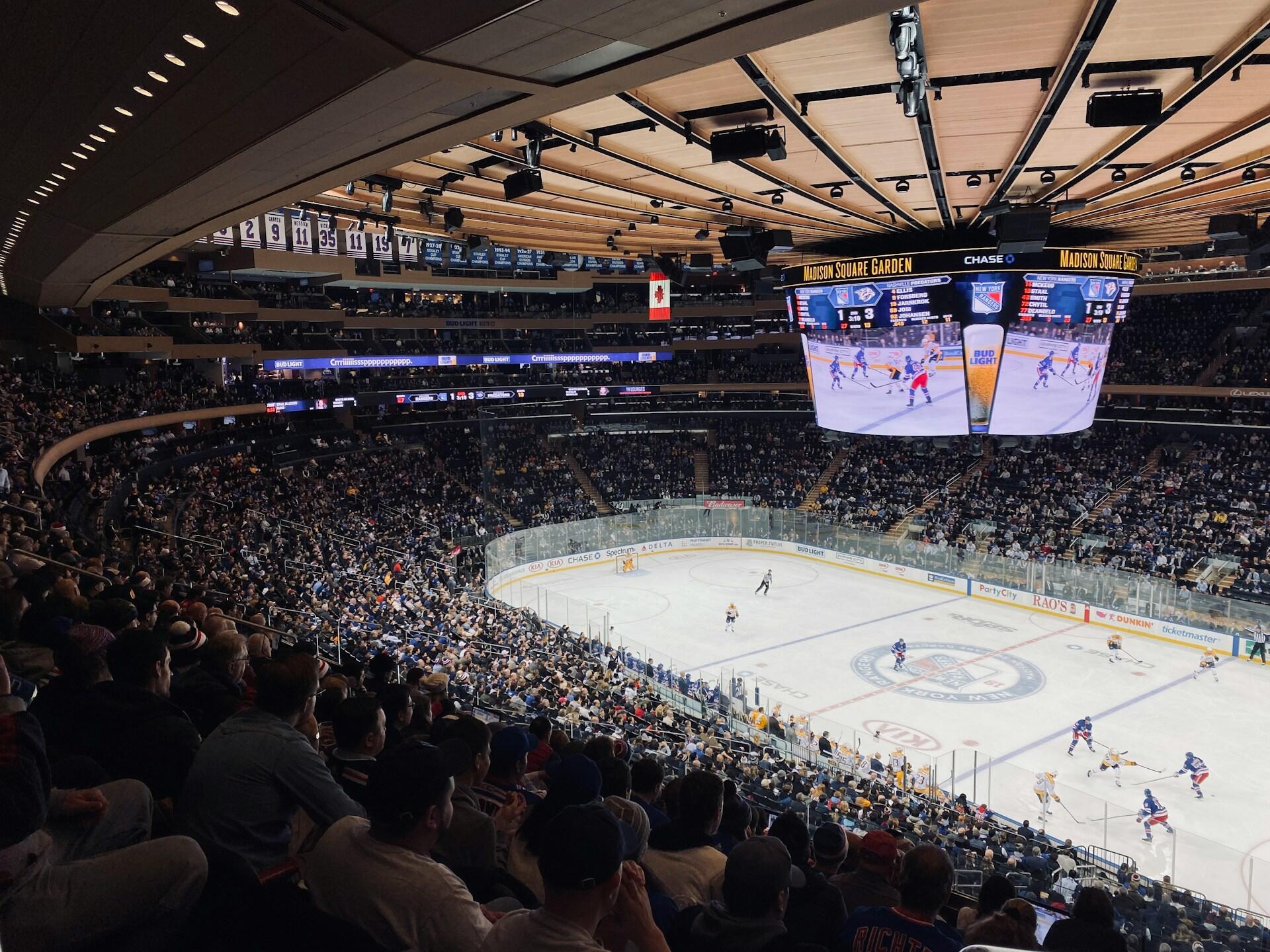 A packed Madison Square Garden during a hockey game, with teams on ice and a large scoreboard displaying the score.
