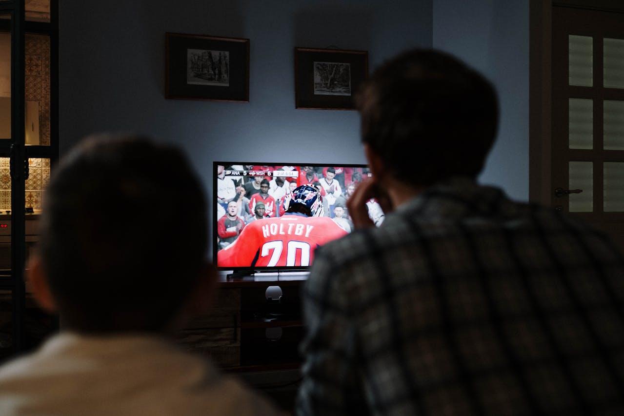 Two people watch a hockey game on TV, focusing on a player in a red jersey with the name "Holtby" on the back.