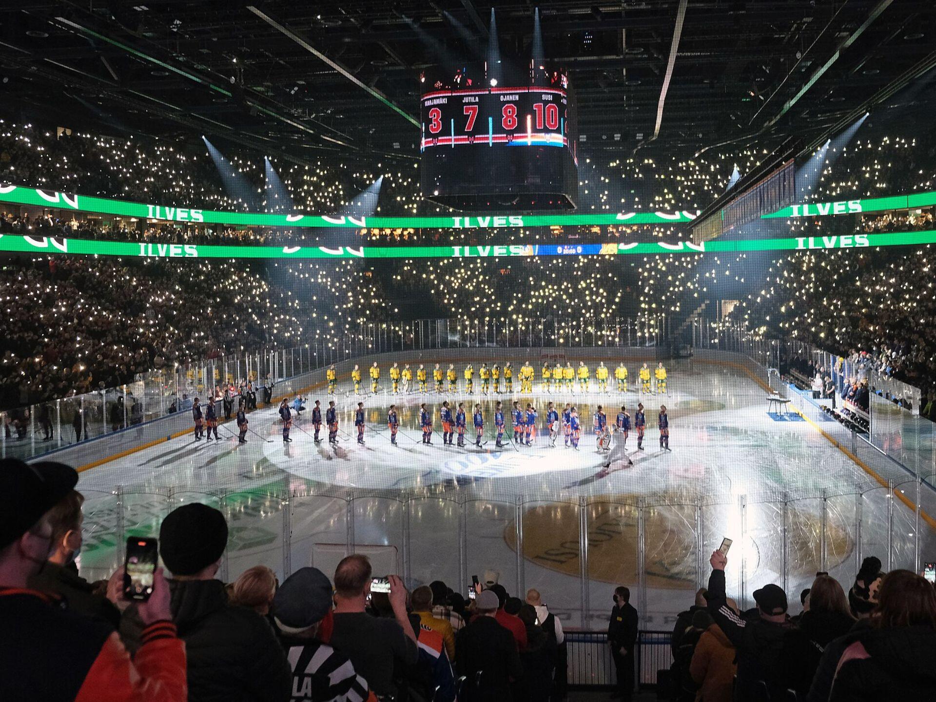 A packed hockey arena with fans using phone lights, teams lined up on the ice, and bright scoreboards overhead amid an electric atmosphere.