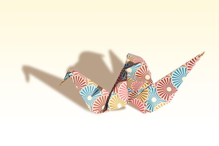 A colorful origami crane with floral patterns casts a soft shadow against a light background.