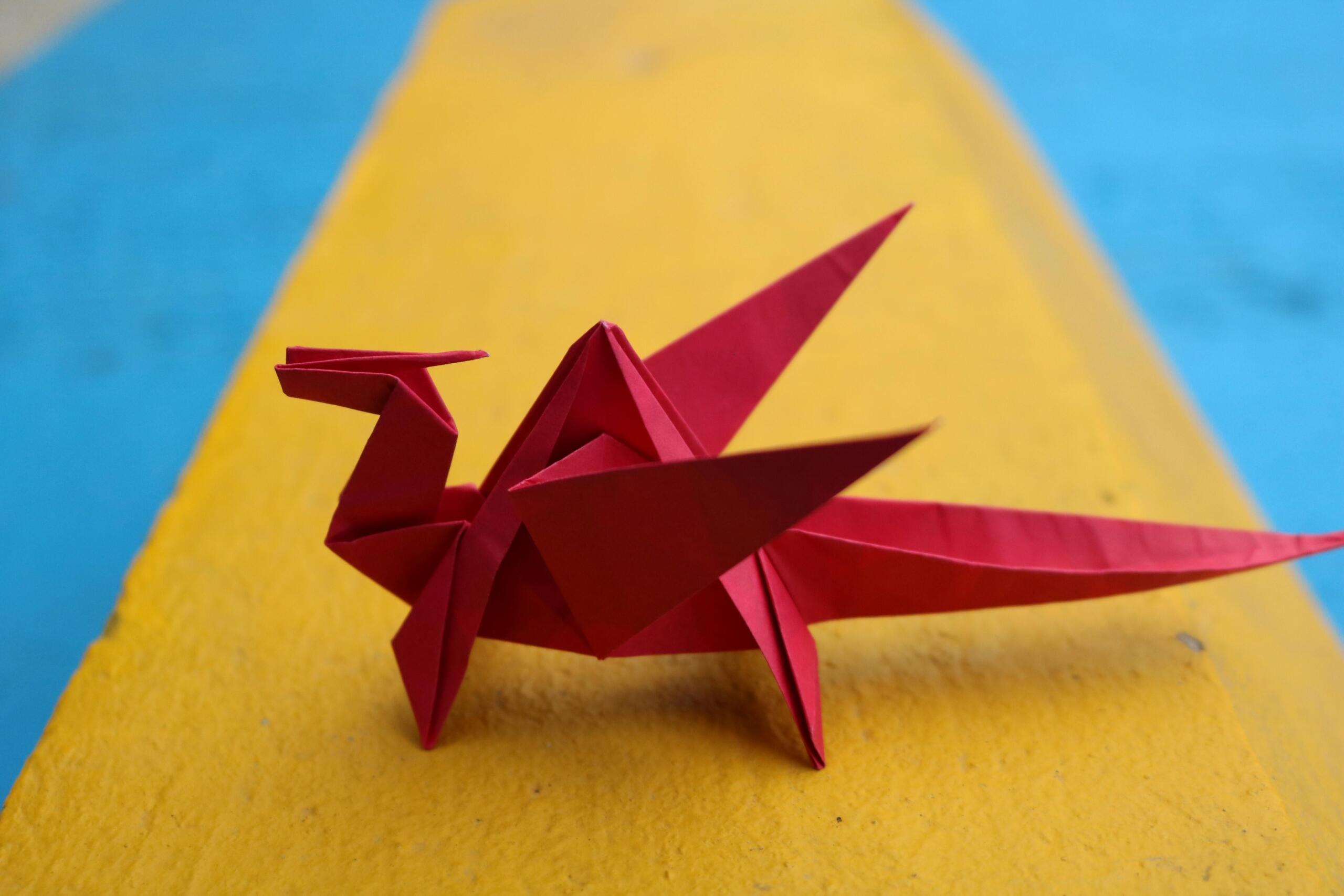 A red origami dragon sits on a vibrant yellow and blue background, showcasing intricate folds and design.