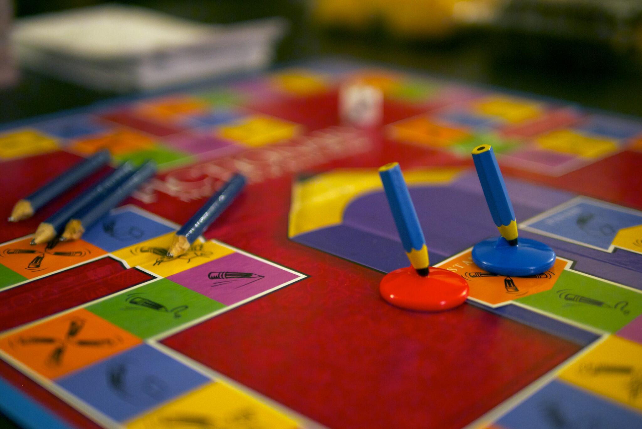 A colorful board game with blue pencils, red and blue game pieces, and various colorful squares showing different symbols and drawings.