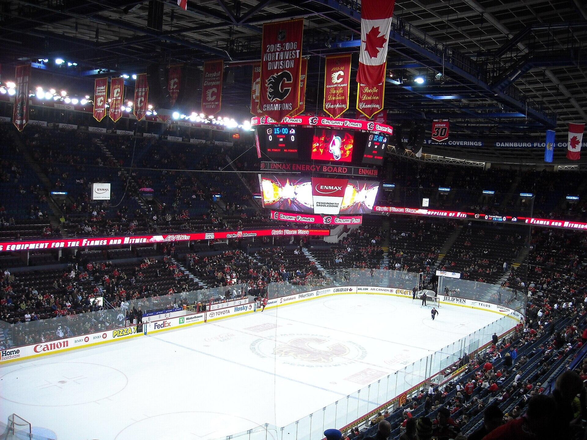 A panoramic view of an ice hockey arena, featuring Calgary Flames banners, a scoreboard, and an excited crowd. The ice rink is prepared for a game.