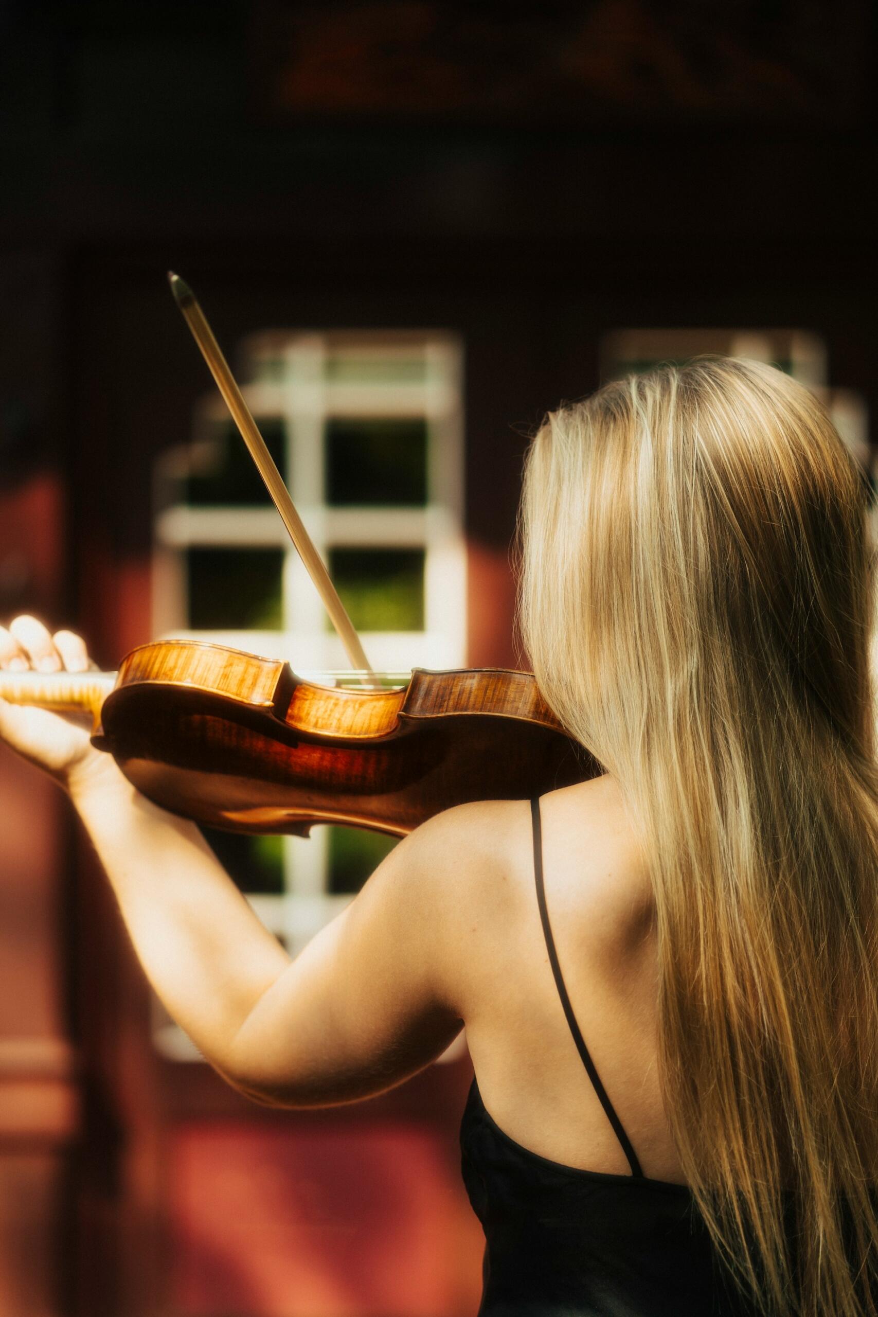 Rear view of a blonde musician playing a violin with a bow in a warmly lit setting.